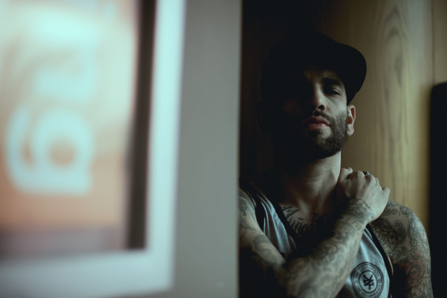 Person with tattoos wearing a baseball cap poses thoughtfully in dramatic lighting.