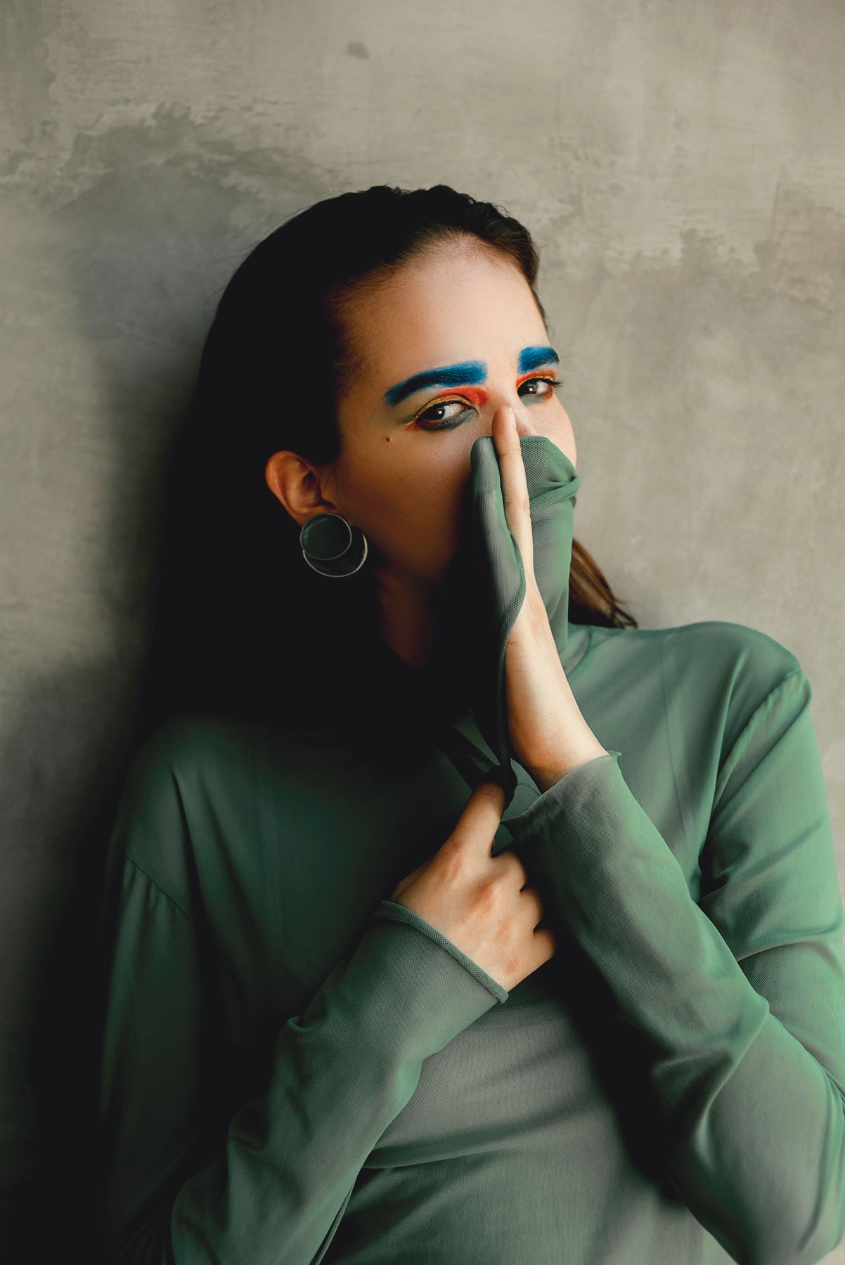 Creative portrait of person with blue eyebrows wearing green sweater against gray wall.