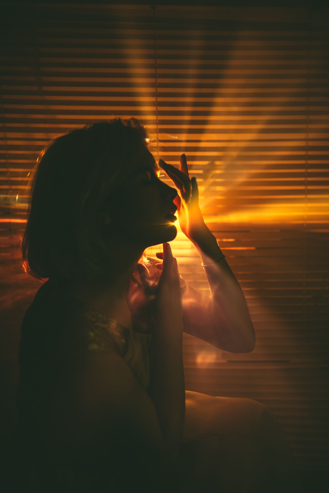 A silhouetted figure stands by window blinds during golden sunset creating dramatic shadows and light beams.