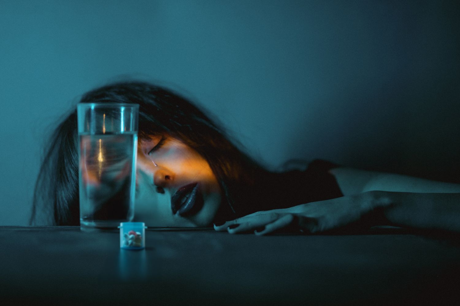 A figure rests beside a glass of water in a moody teal-tinted scene, creating a dramatic atmospheric composition.