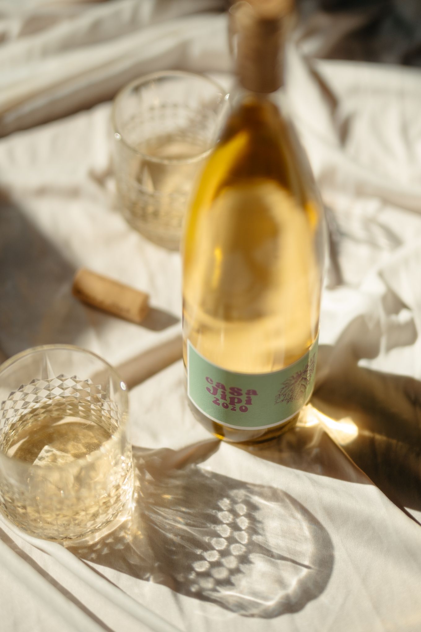 A wine bottle and crystal glass casting dramatic shadows on white fabric in warm sunlight.