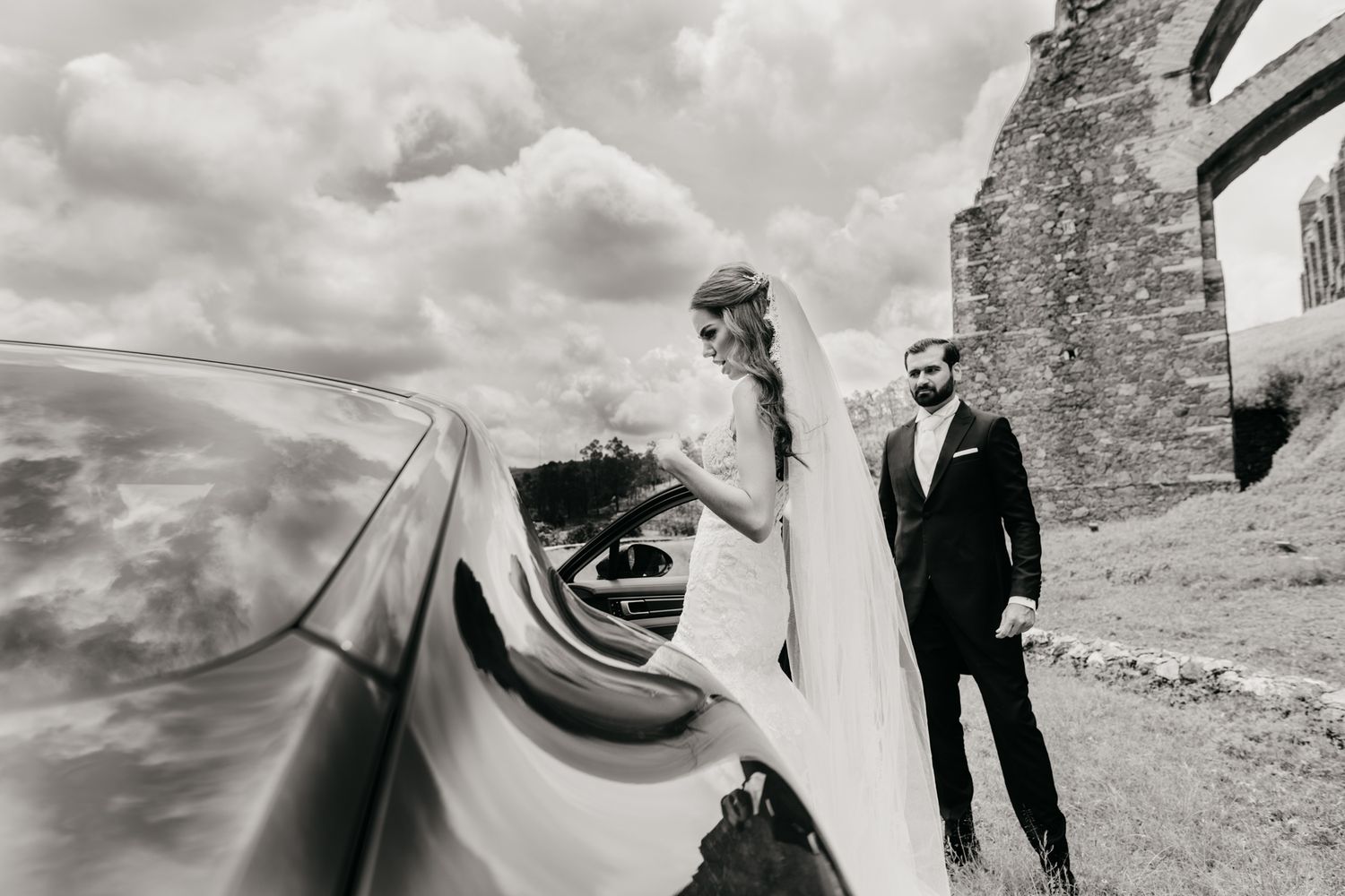 A black and white wedding photo of a couple near a classic car and stone building in an artistic composition.