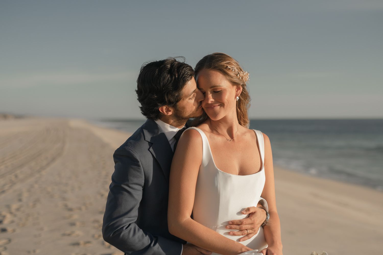 A romantic couple embraces on a serene beach at sunset with the ocean waves in the background.