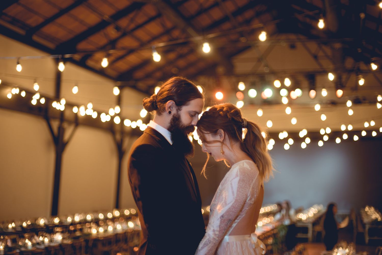 A romantic wedding couple shares an intimate moment underneath warm string lights in a rustic indoor venue.
