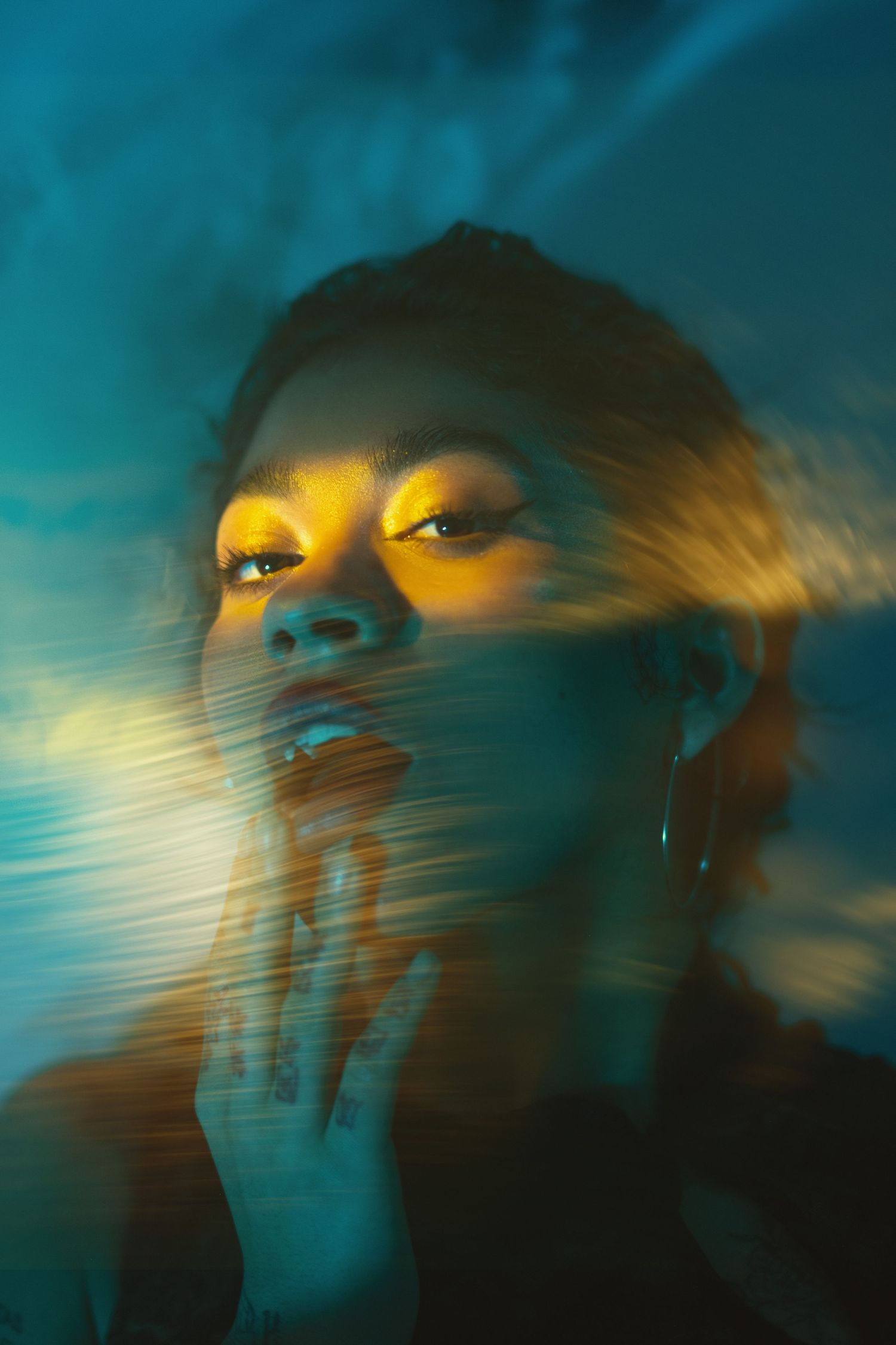 Artistic portrait with dramatic teal and yellow lighting creates ethereal motion blur effect against dark background.
