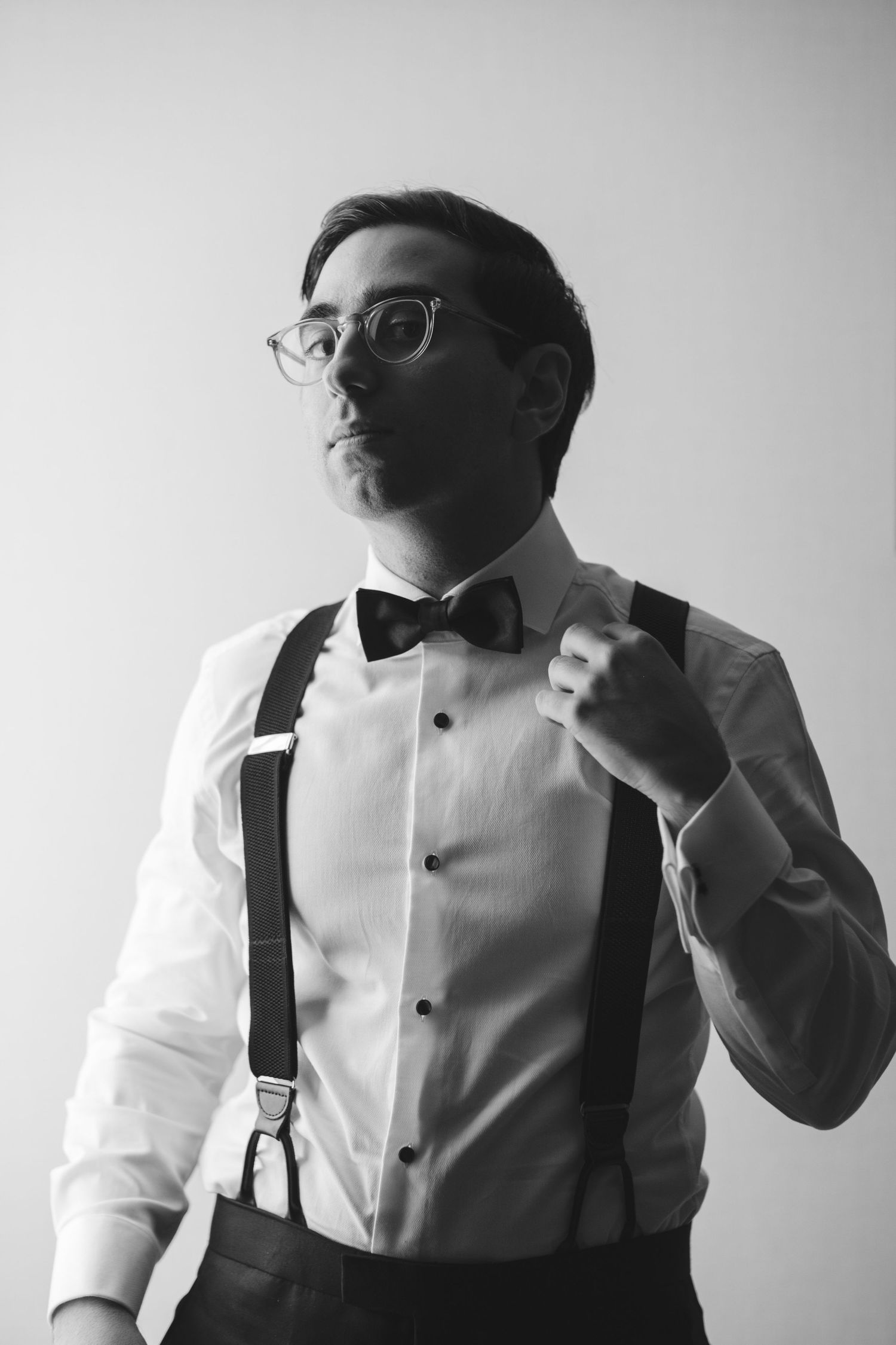 Black and white portrait of groom adjusting bowtie while wearing suspenders and glasses.