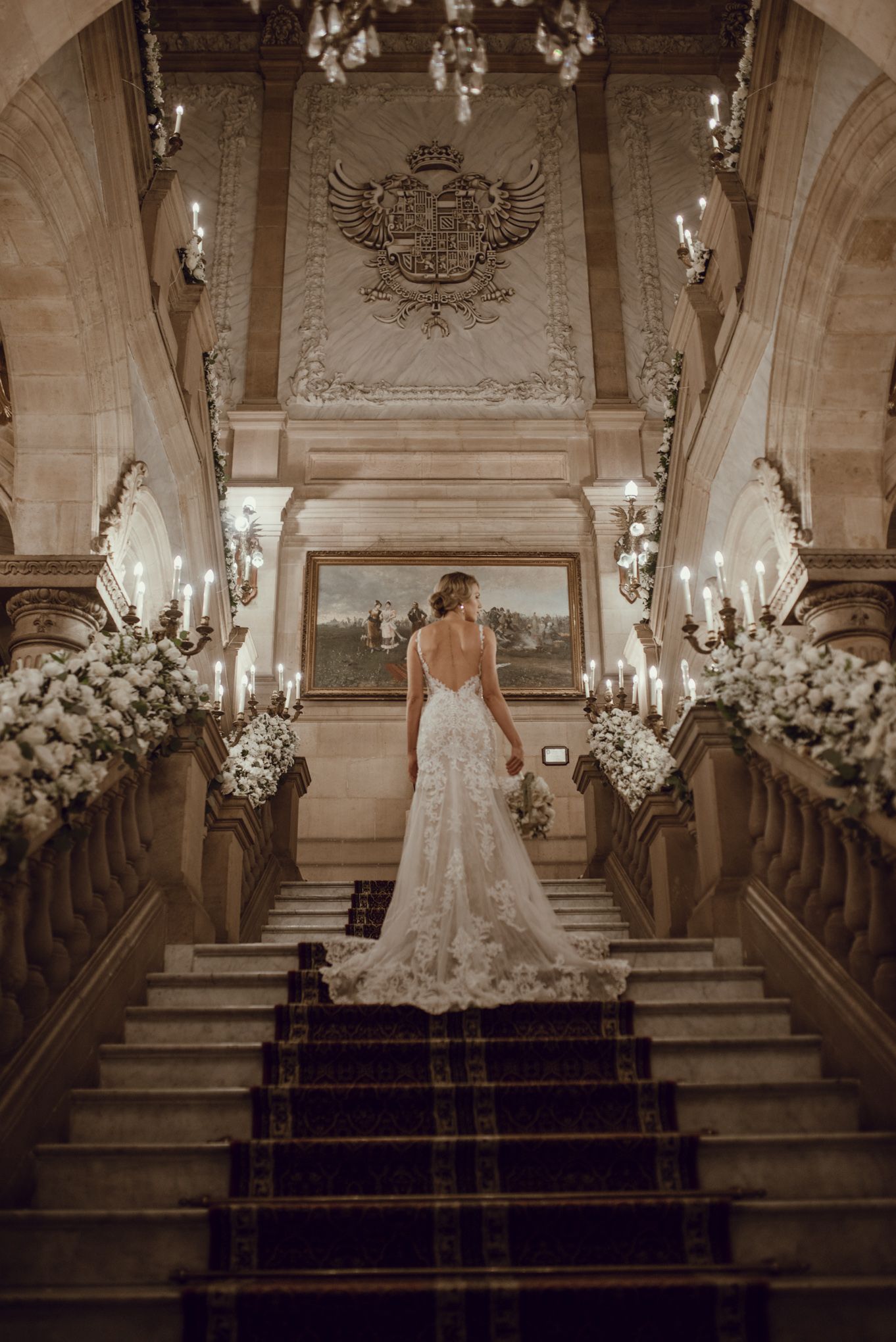 A bride in a lace wedding gown stands on a grand staircase adorned with floral arrangements and chandeliers.