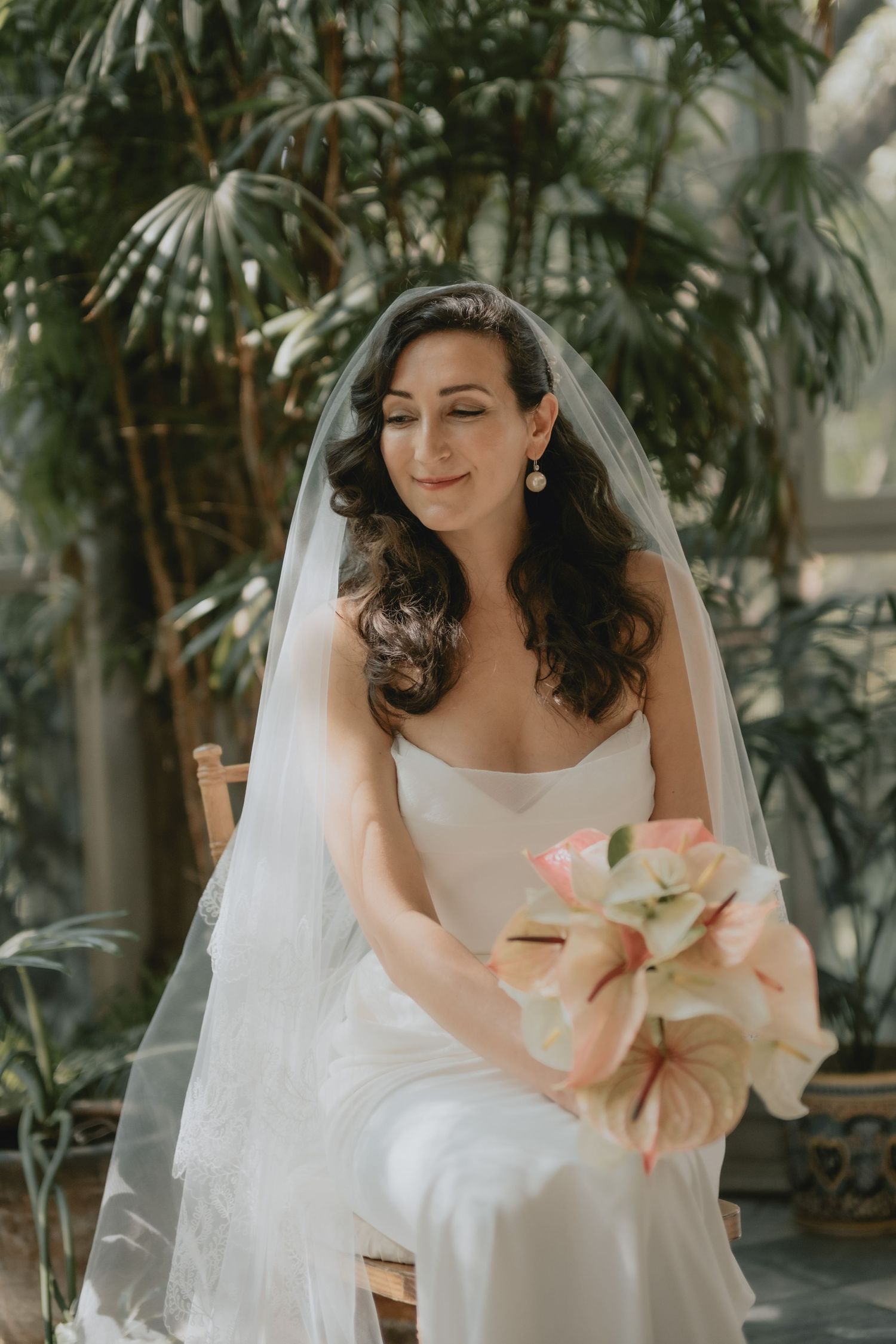 A bride in a white gown holds a pink bouquet while seated in a greenhouse setting.