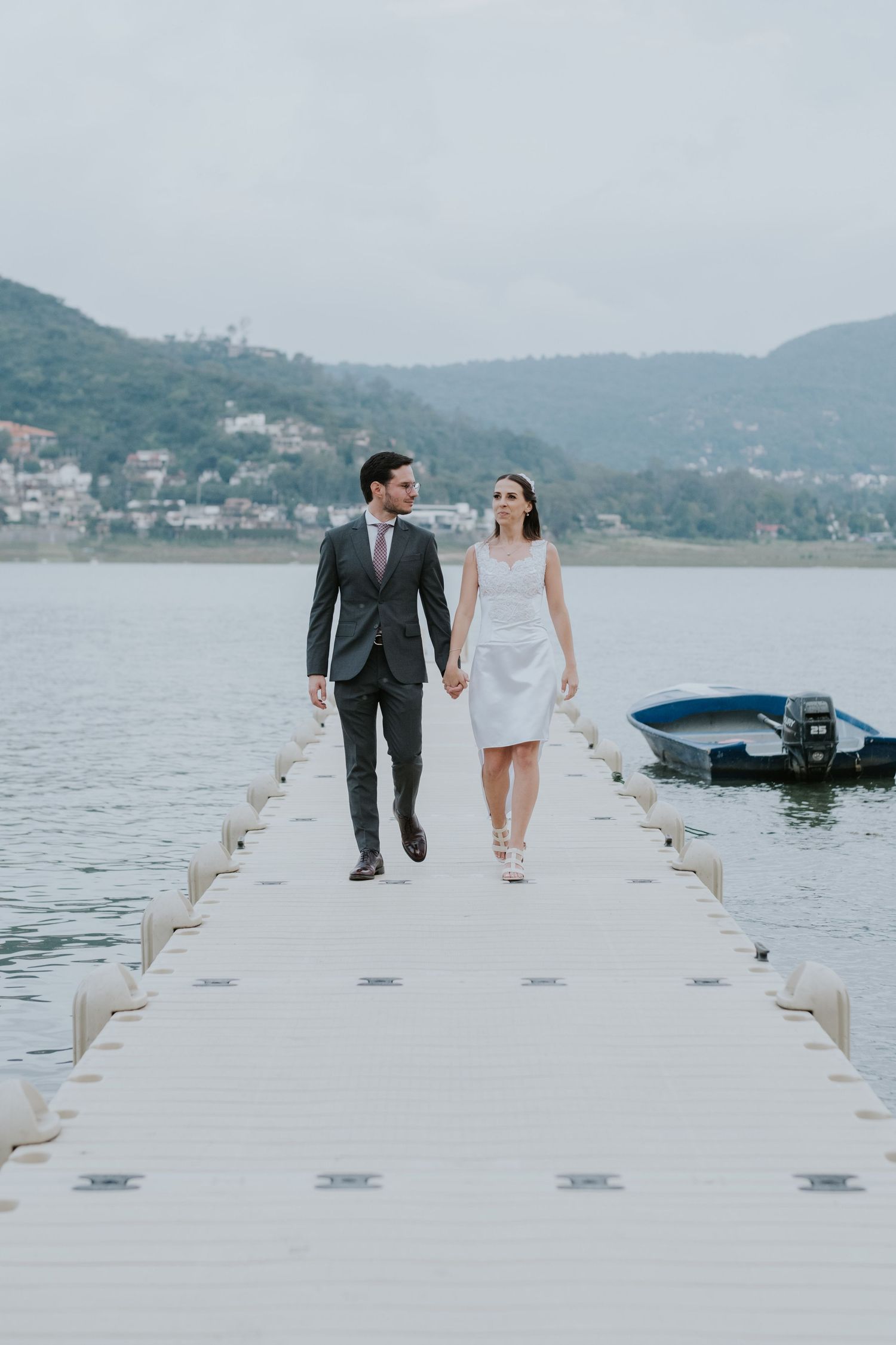 Couple walking hand in hand on white pier over serene lake.