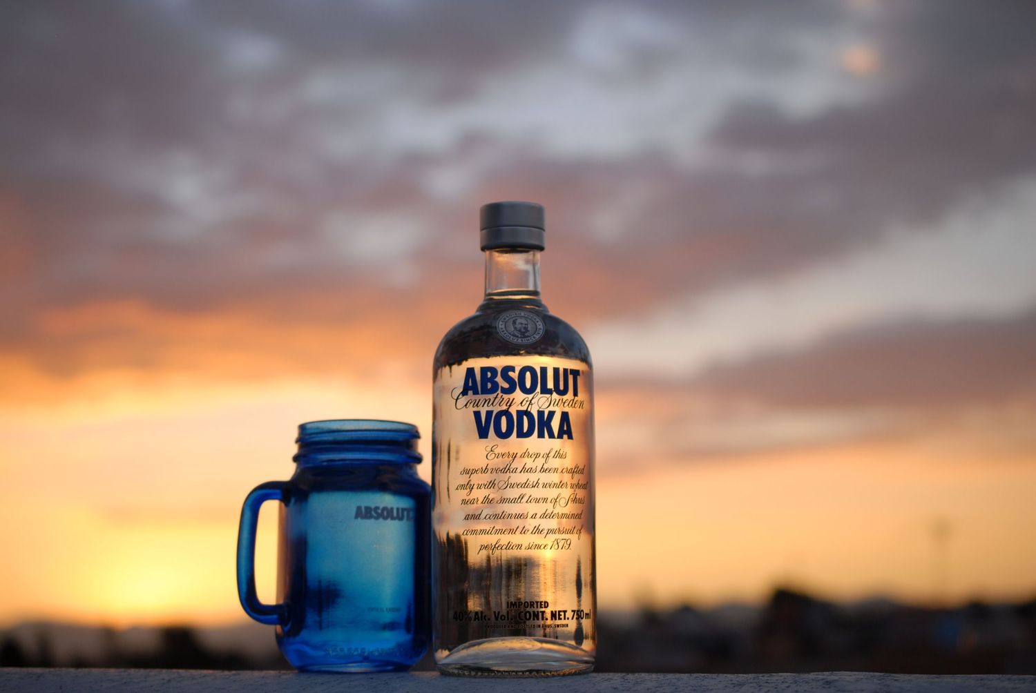 Absolut Vodka bottle and blue mason jar silhouetted against sunset sky.