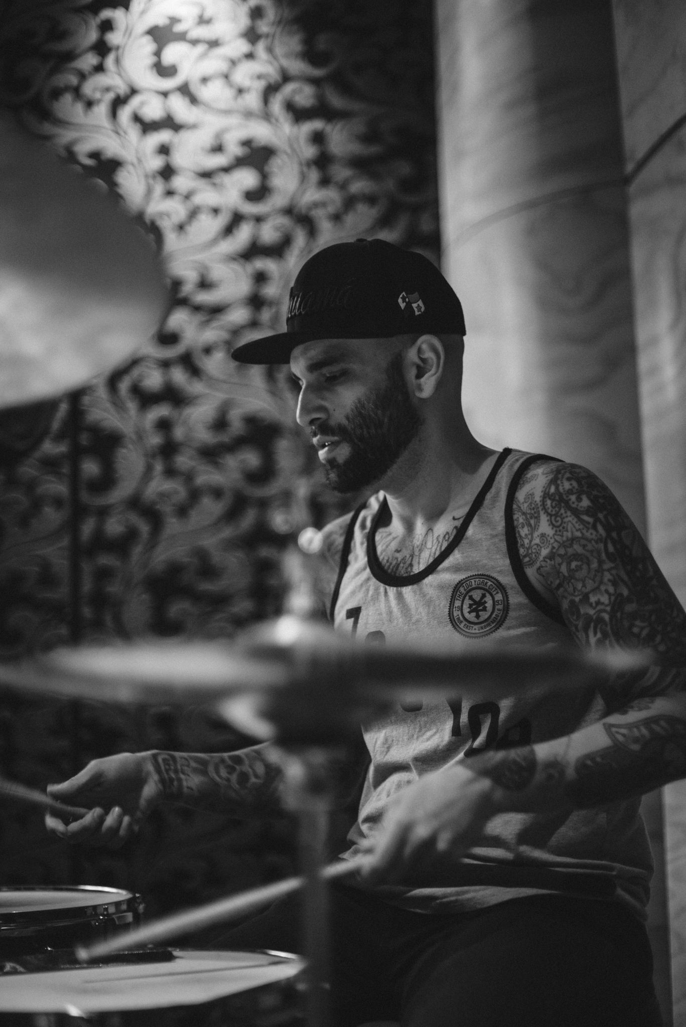 Black and white photo of a drummer with tattoos performing on stage with dramatic side lighting.