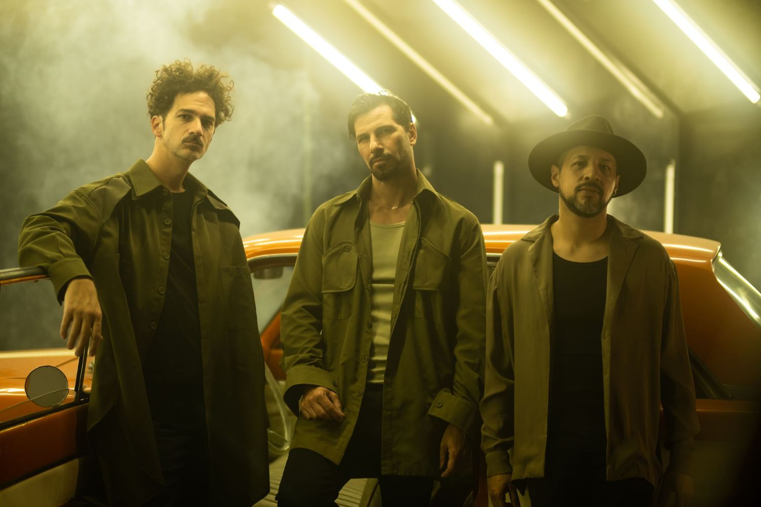 Three performers stand in moody yellow lighting with atmospheric fog effects.