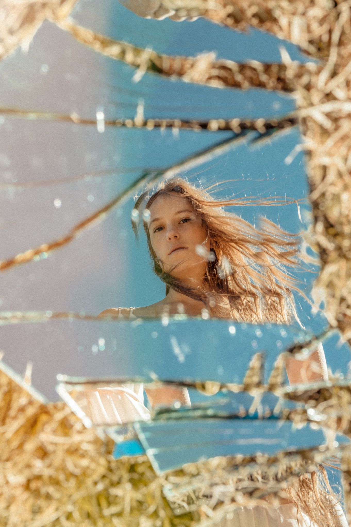 Artistic portrait through broken mirror fragments reflecting blue sky and windswept hair on a sunny day.