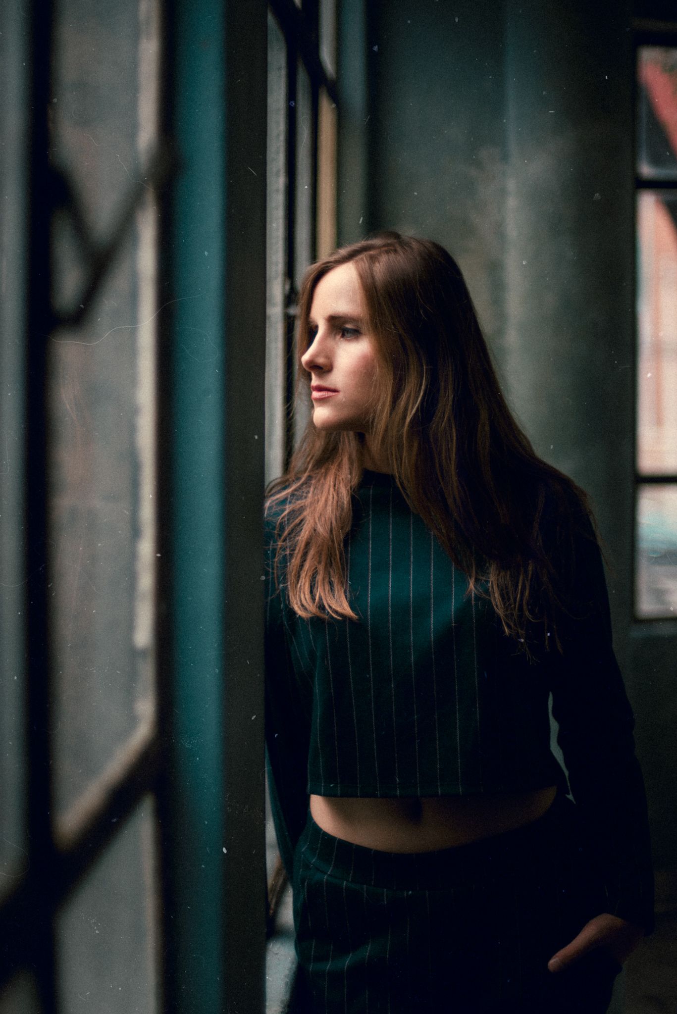 A person in a dark striped outfit looks pensively through a window in moody lighting.
