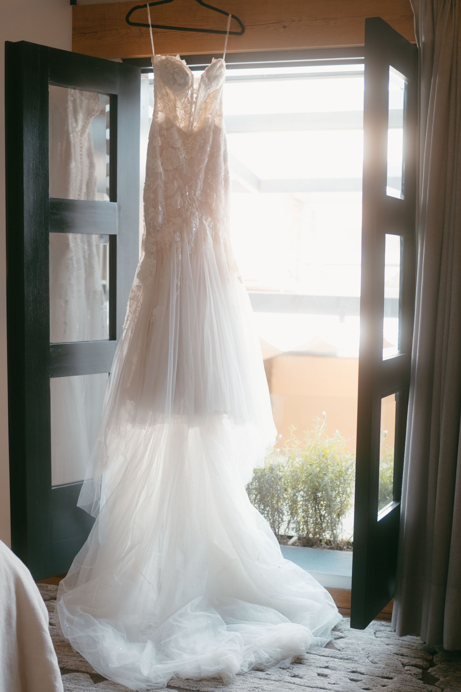 Elegant white wedding dress with lace detailing hanging in window light against dark doors creating romantic ambiance.