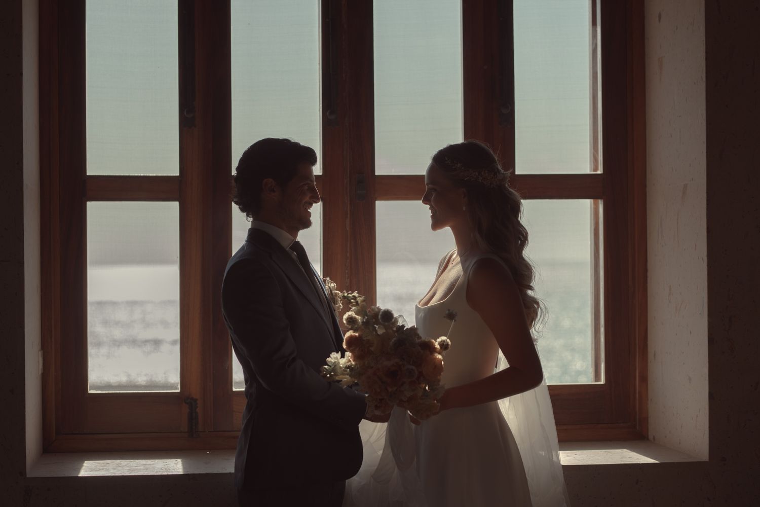 Silhouetted couple stands by large window sharing intimate moment with flowers between them in moody lighting.