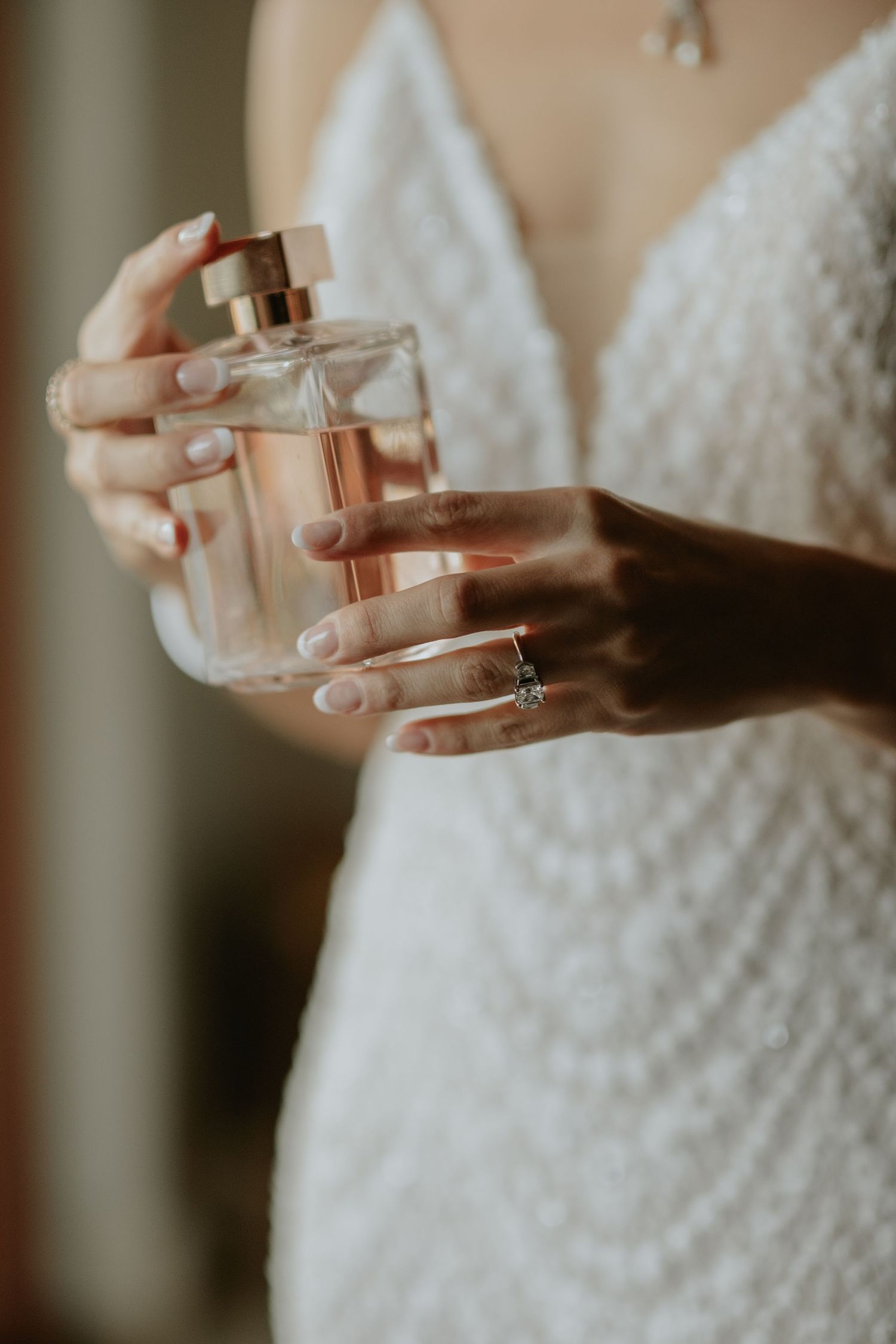 Close-up of hands holding a perfume bottle while wearing a white lace dress and engagement ring.
