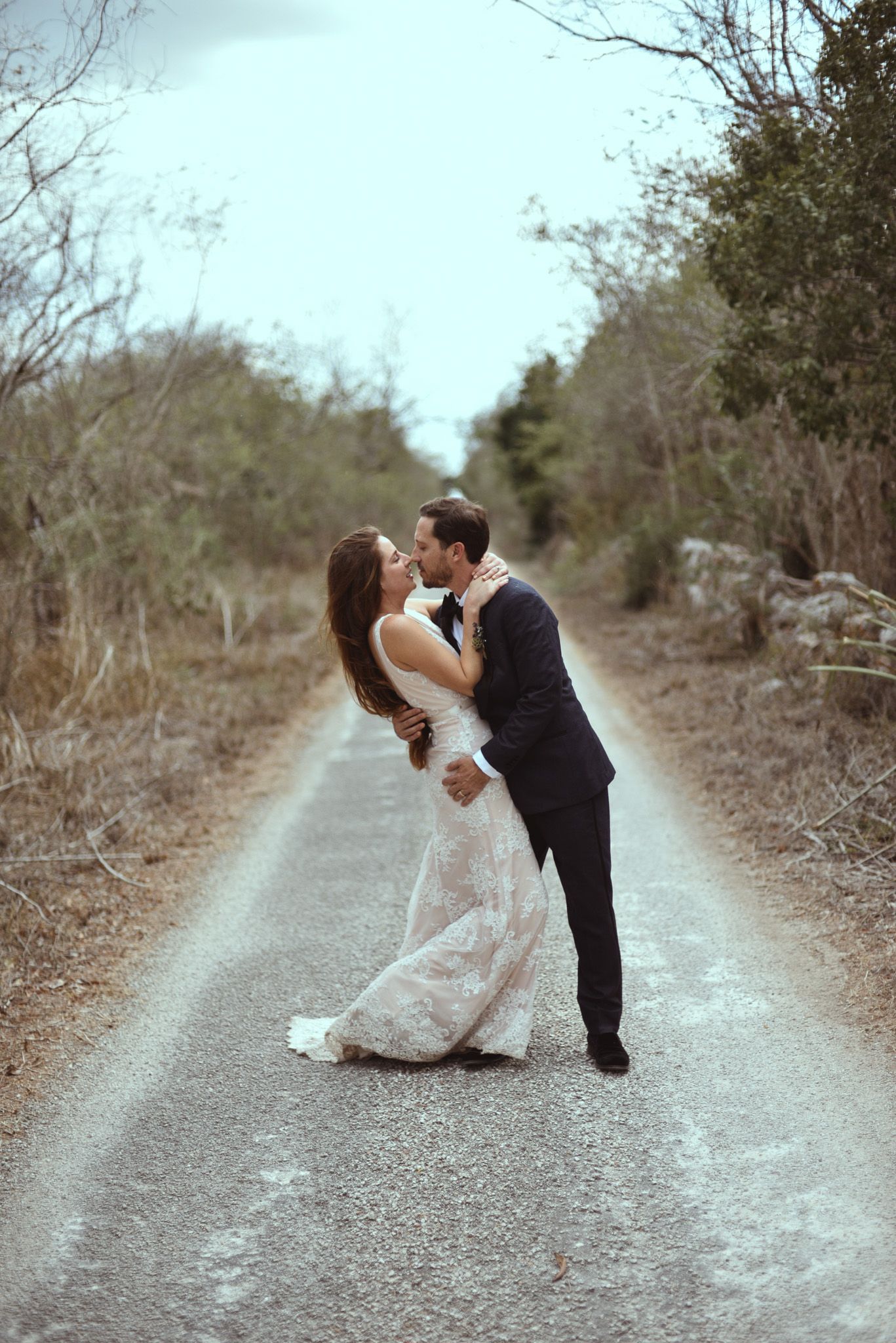 A romantic moment on a rural path captures two figures in formal attire sharing a loving embrace against a blurred forest backdrop.