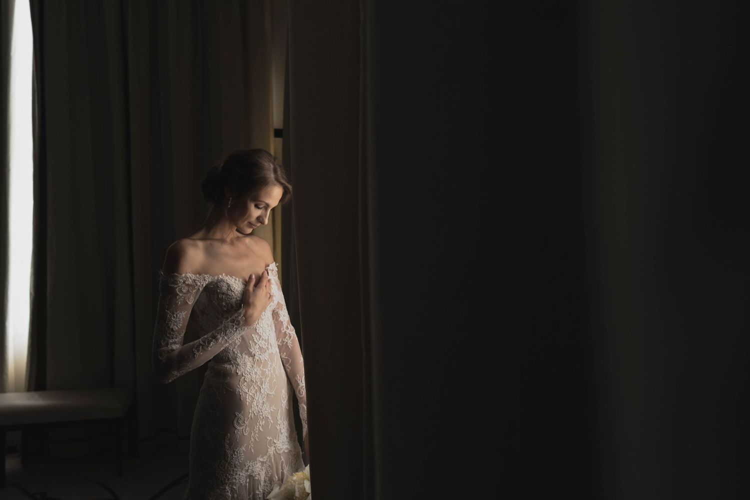 A figure in a white lace wedding dress stands near a window in moody lighting.