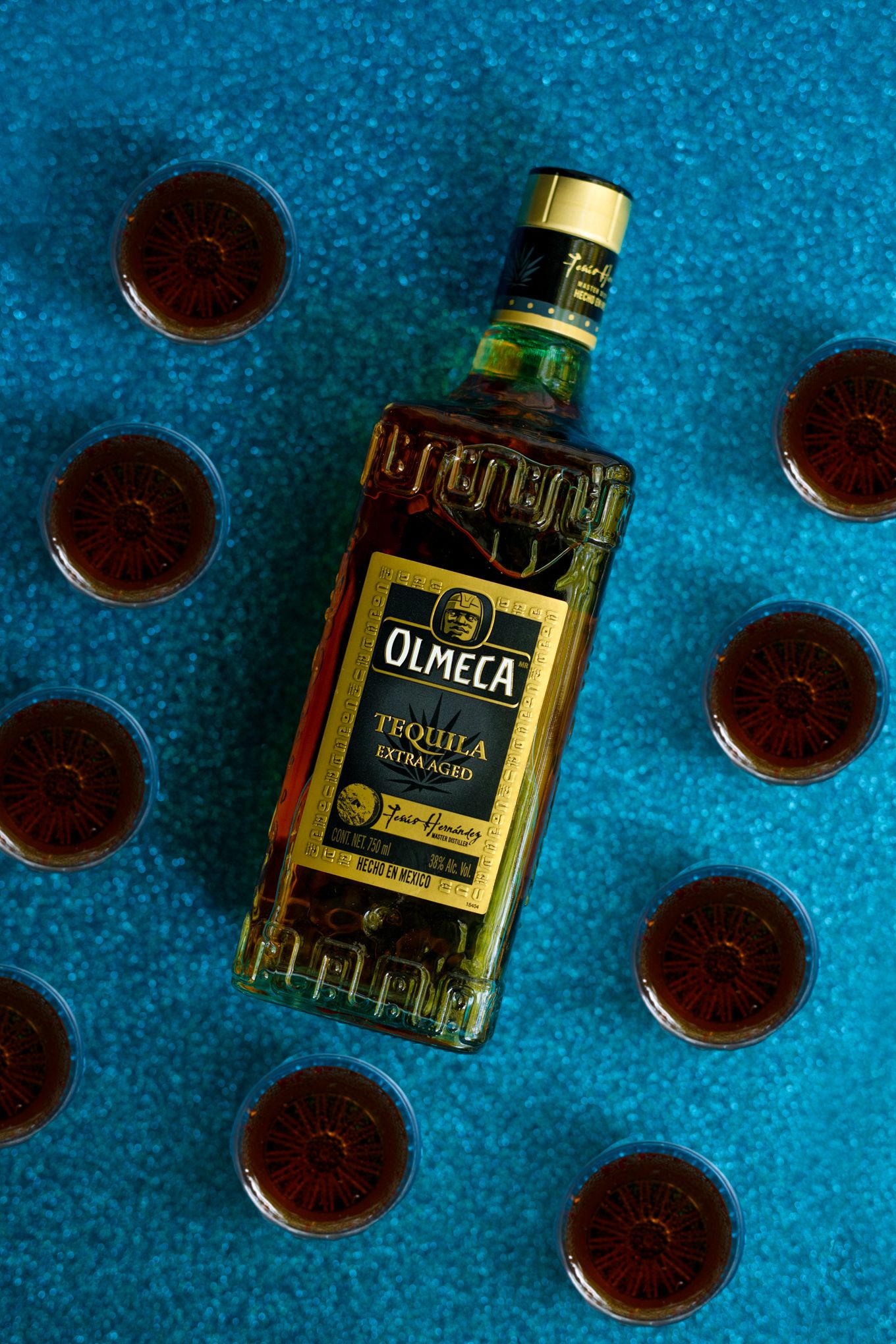 Overhead view of Olmeca tequila bottle surrounded by filled shot glasses on blue background.