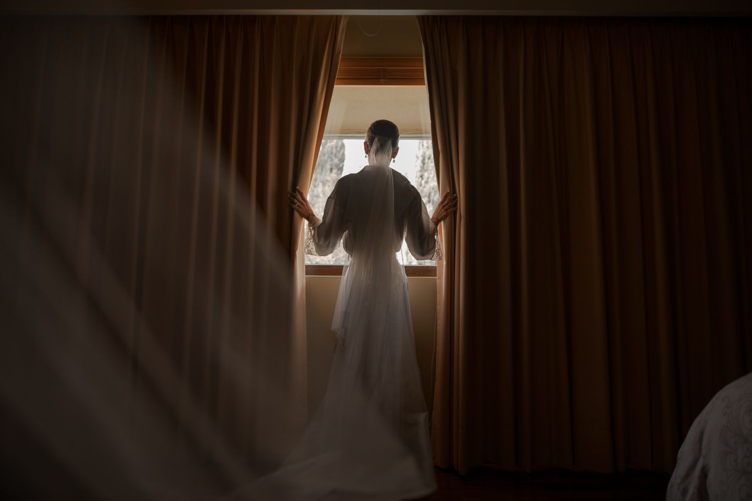 Silhouette of bride in wedding dress standing at window with dramatic curtains.