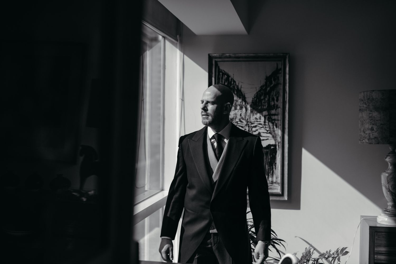 Sophisticated figure in black suit gazes thoughtfully through bright window in moody black and white portrait.