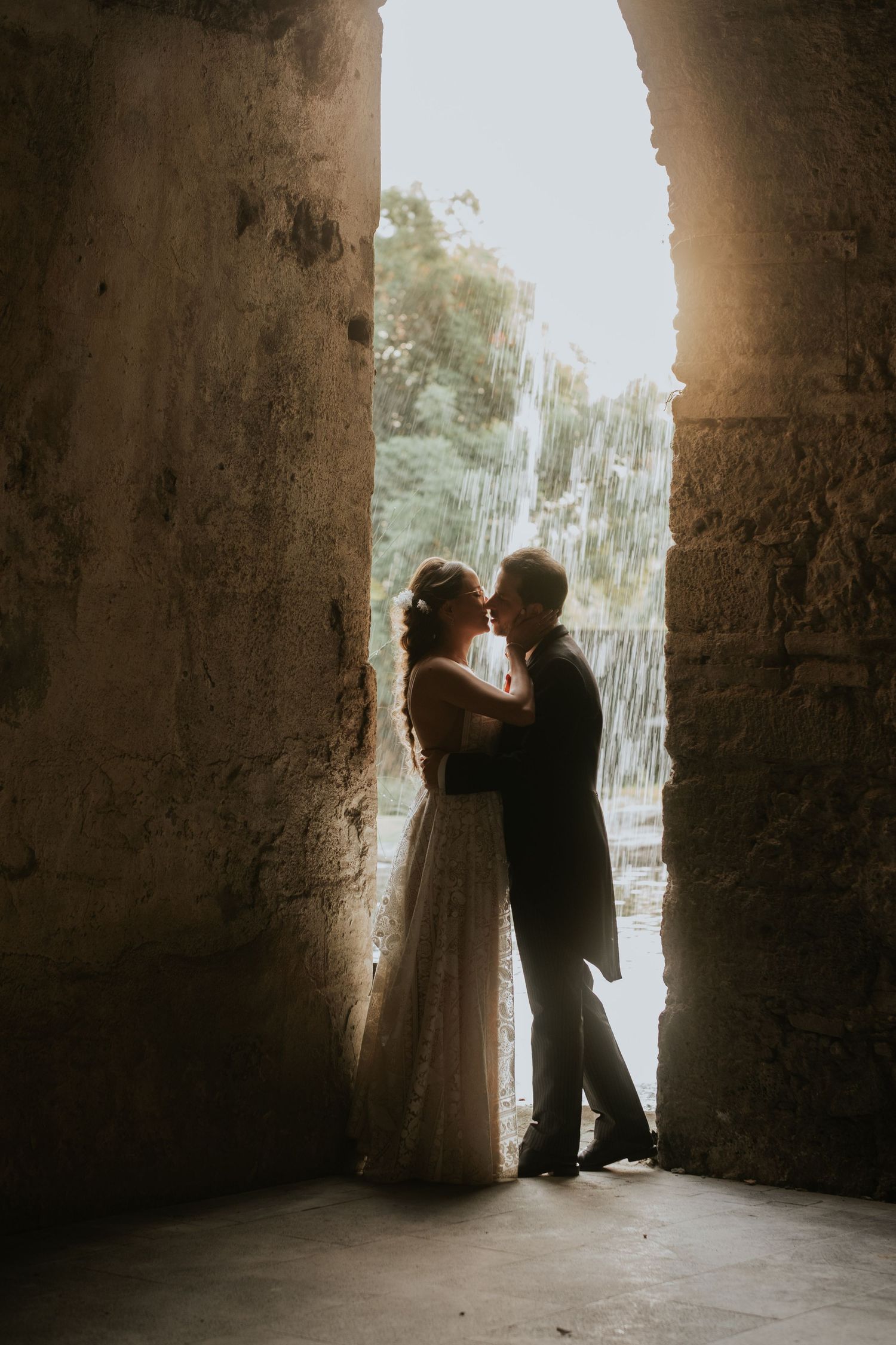 A silhouetted wedding couple embraces in a stone archway with backlit natural light.