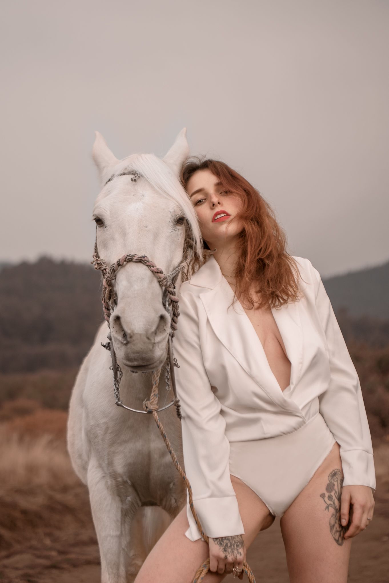 A white horse and person in white bodysuit and blazer pose together in a moody outdoor setting.