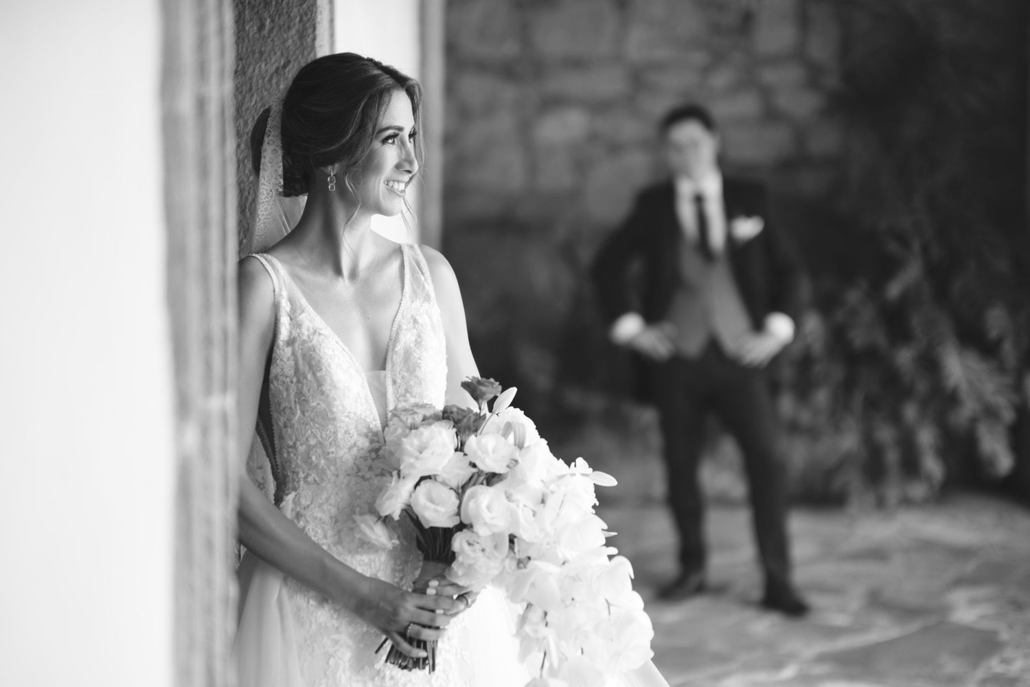 Bride holding white bouquet poses in elegant lace dress while groom stands in background.