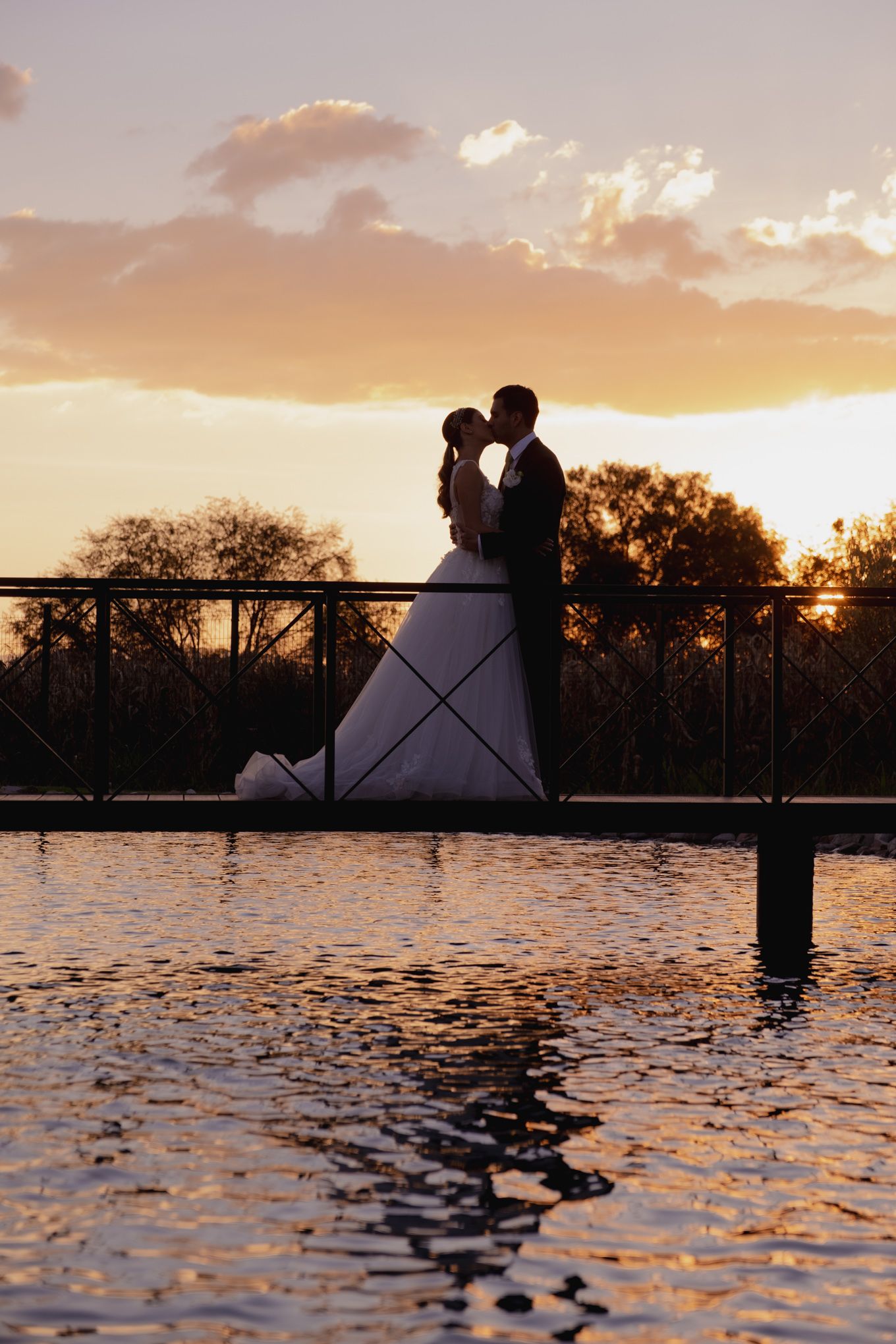 Silhouetted couple shares romantic moment on bridge during golden sunset over water.