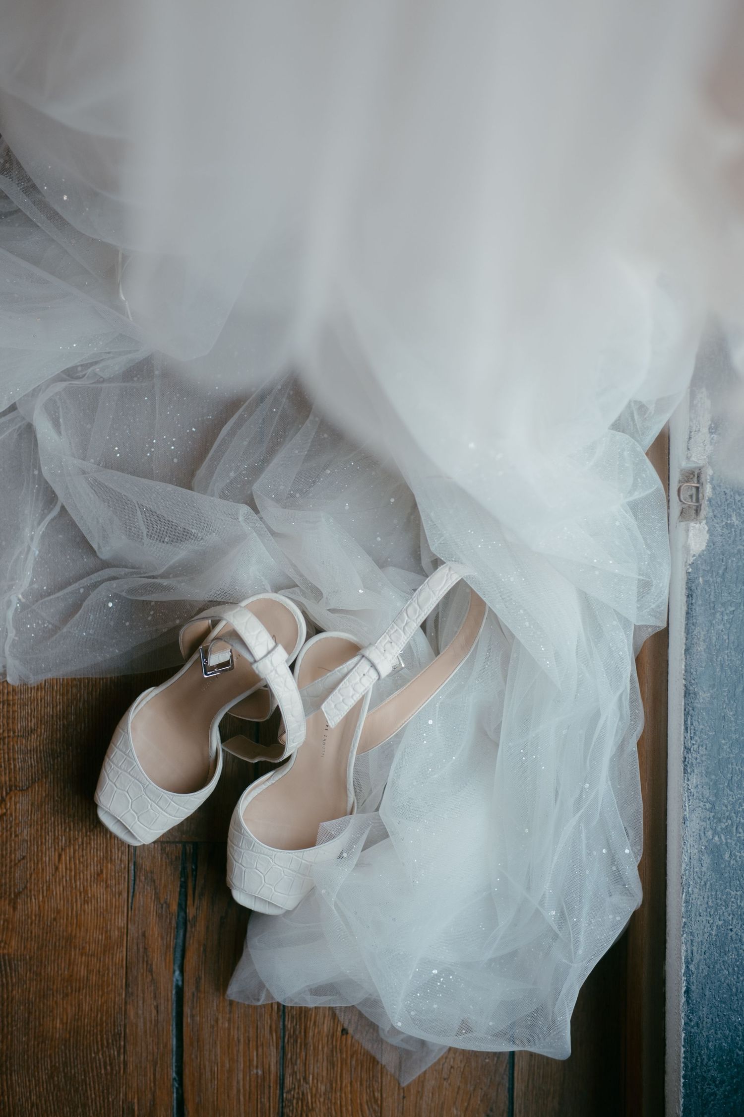 White wedding shoes nestled among flowing tulle fabric on wooden floor.