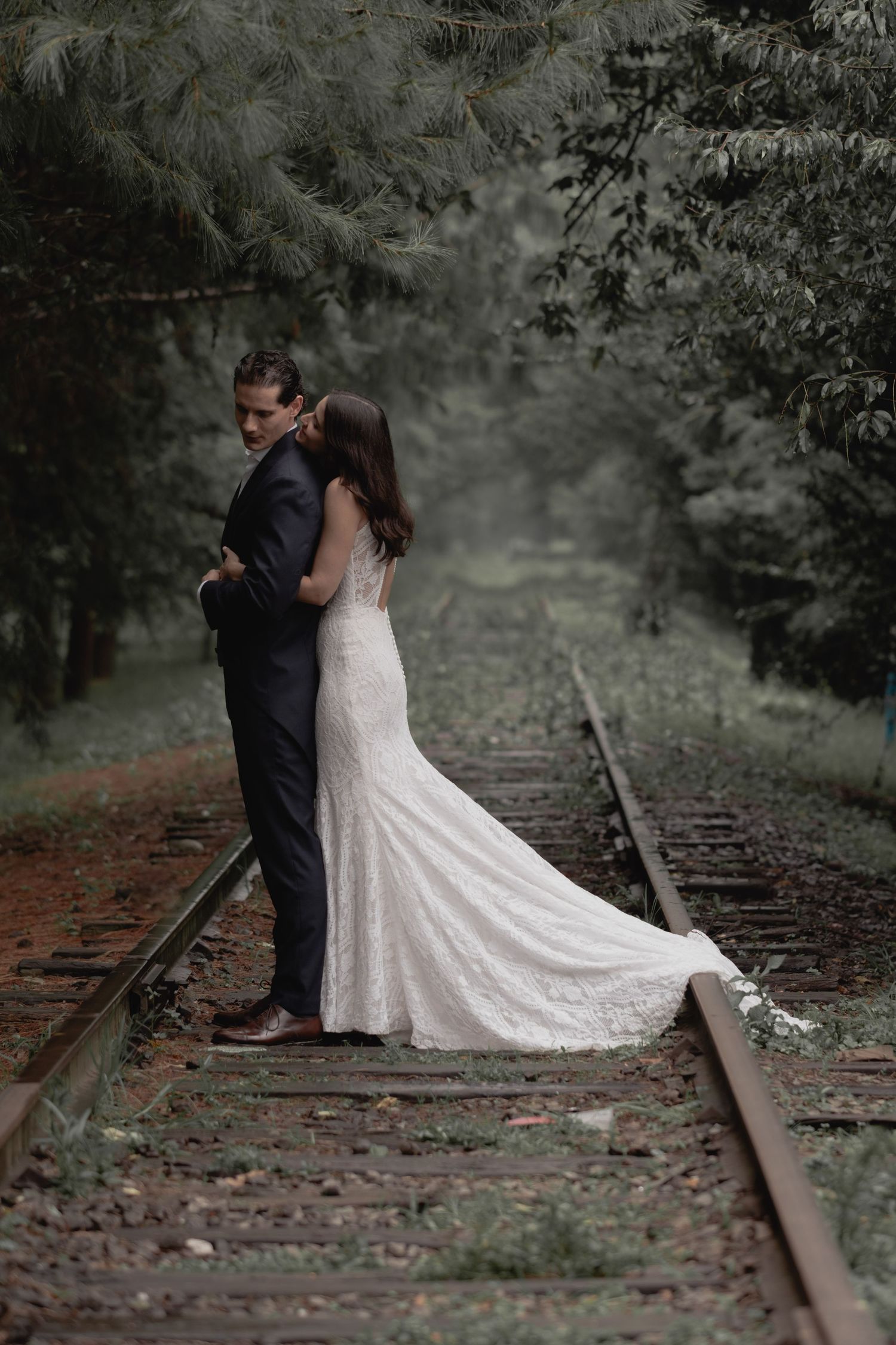 A romantic wedding portrait of a couple embracing on rustic train tracks surrounded by forest.