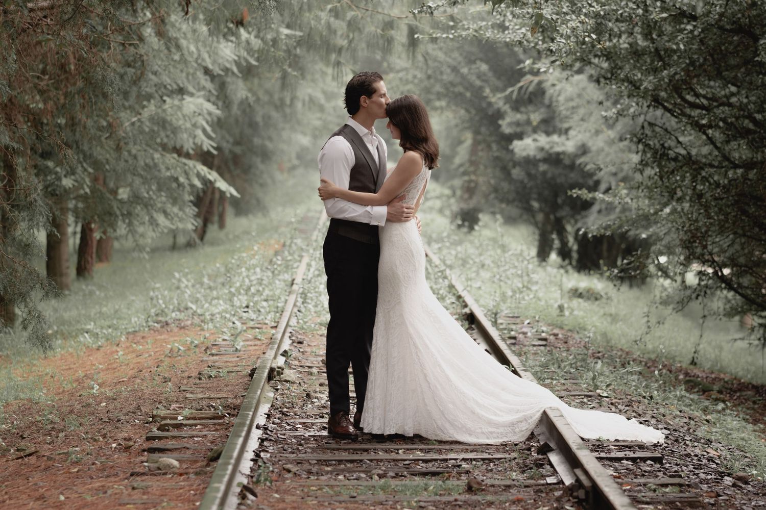 A romantic wedding couple embraces on rustic train tracks surrounded by misty forest scenery.