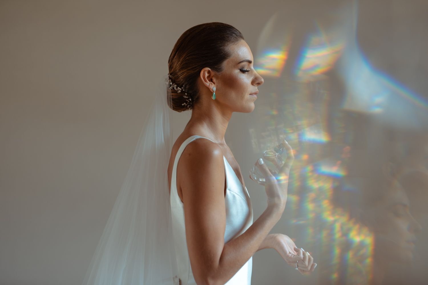 Bride in elegant white gown stands in soft light with rainbow prism reflections.