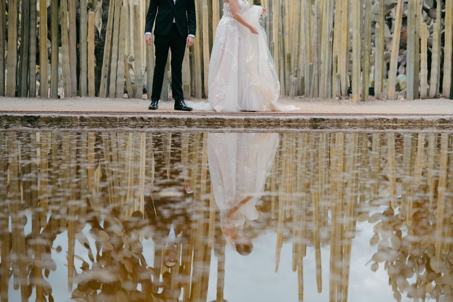 Artistic wedding photo showing couple's reflection in pool by bamboo fence.