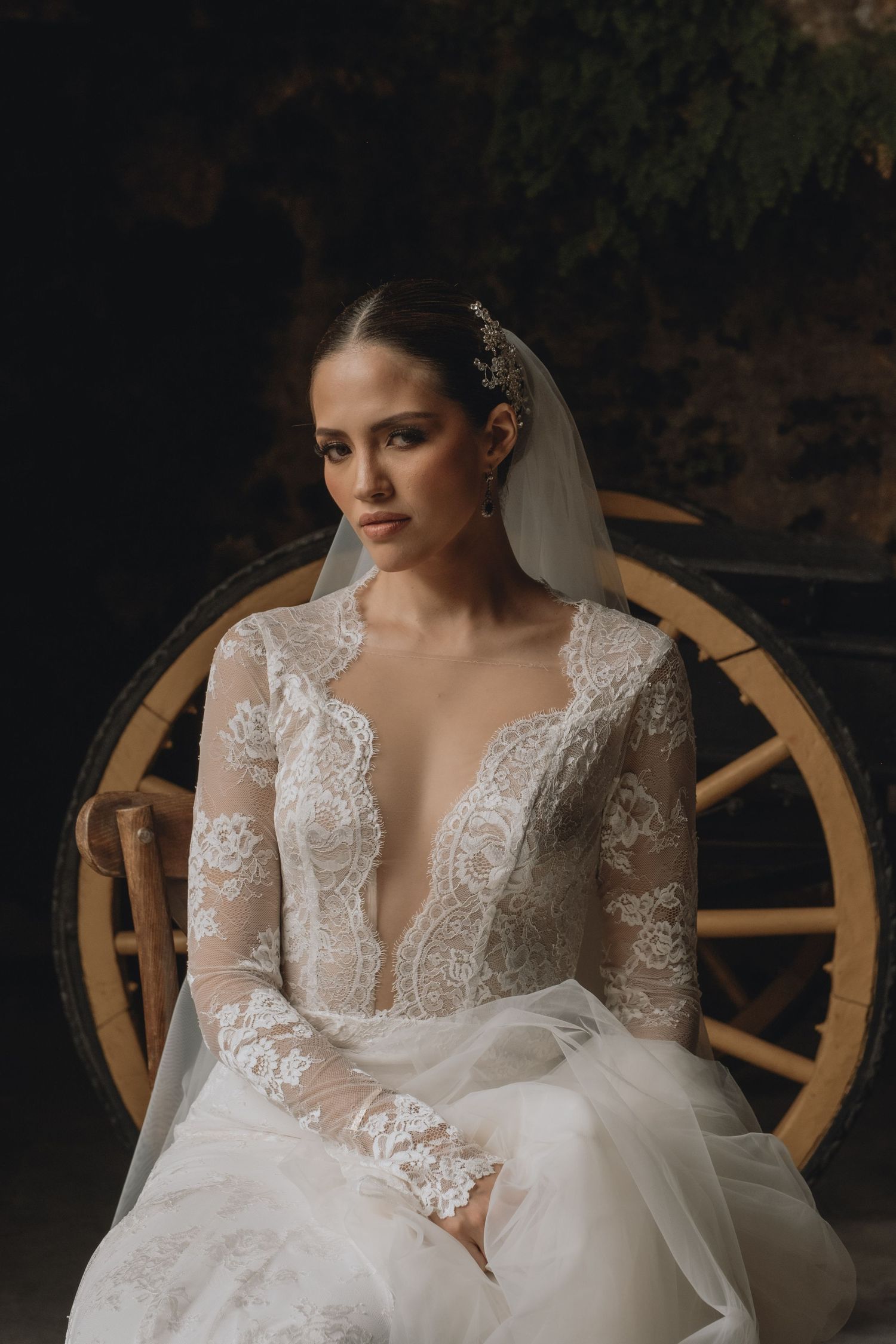 A bride in an elegant lace wedding dress sits against a wooden wheel backdrop.