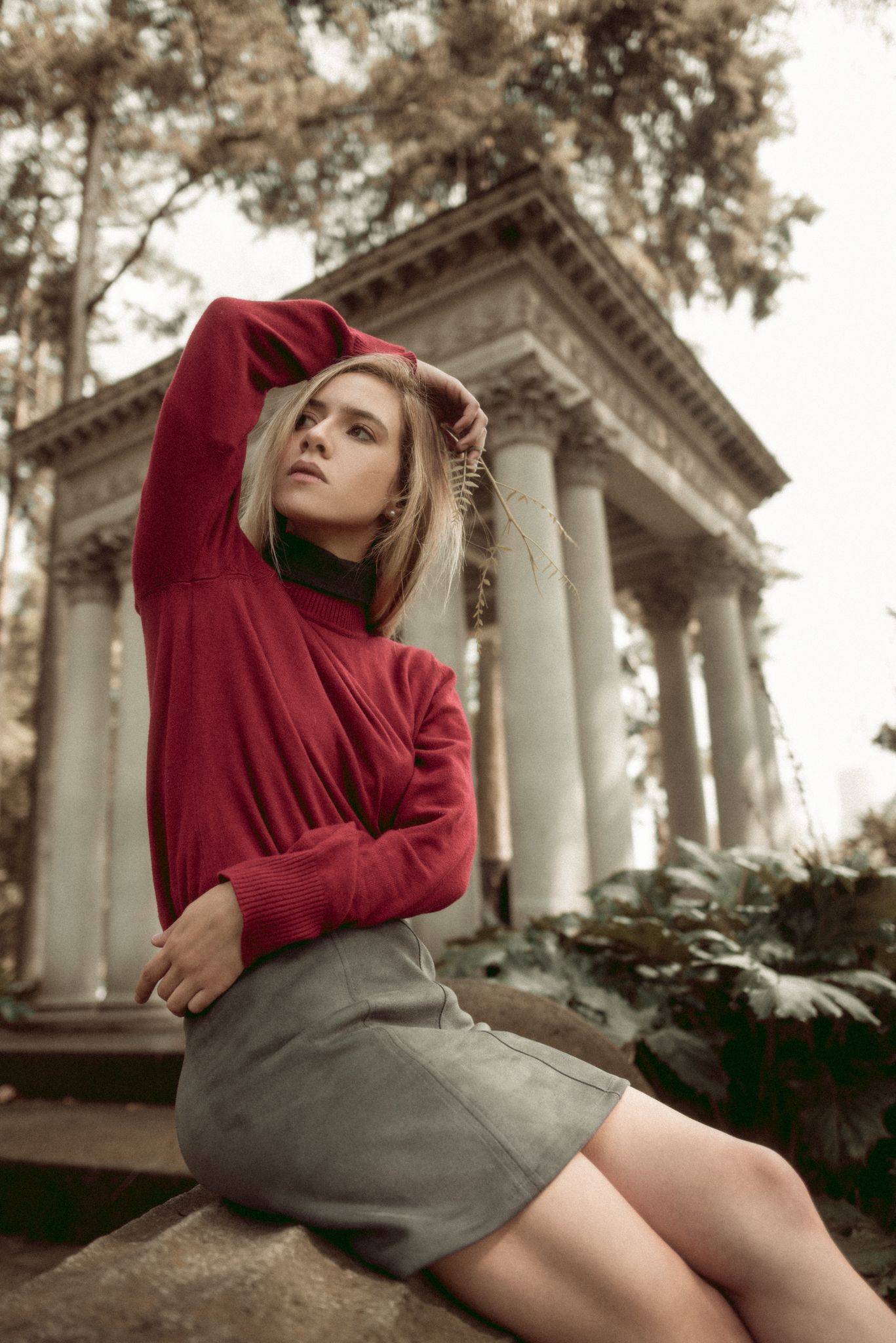 Stylish portrait of person in red sweater and gray skirt against classical columns backdrop.