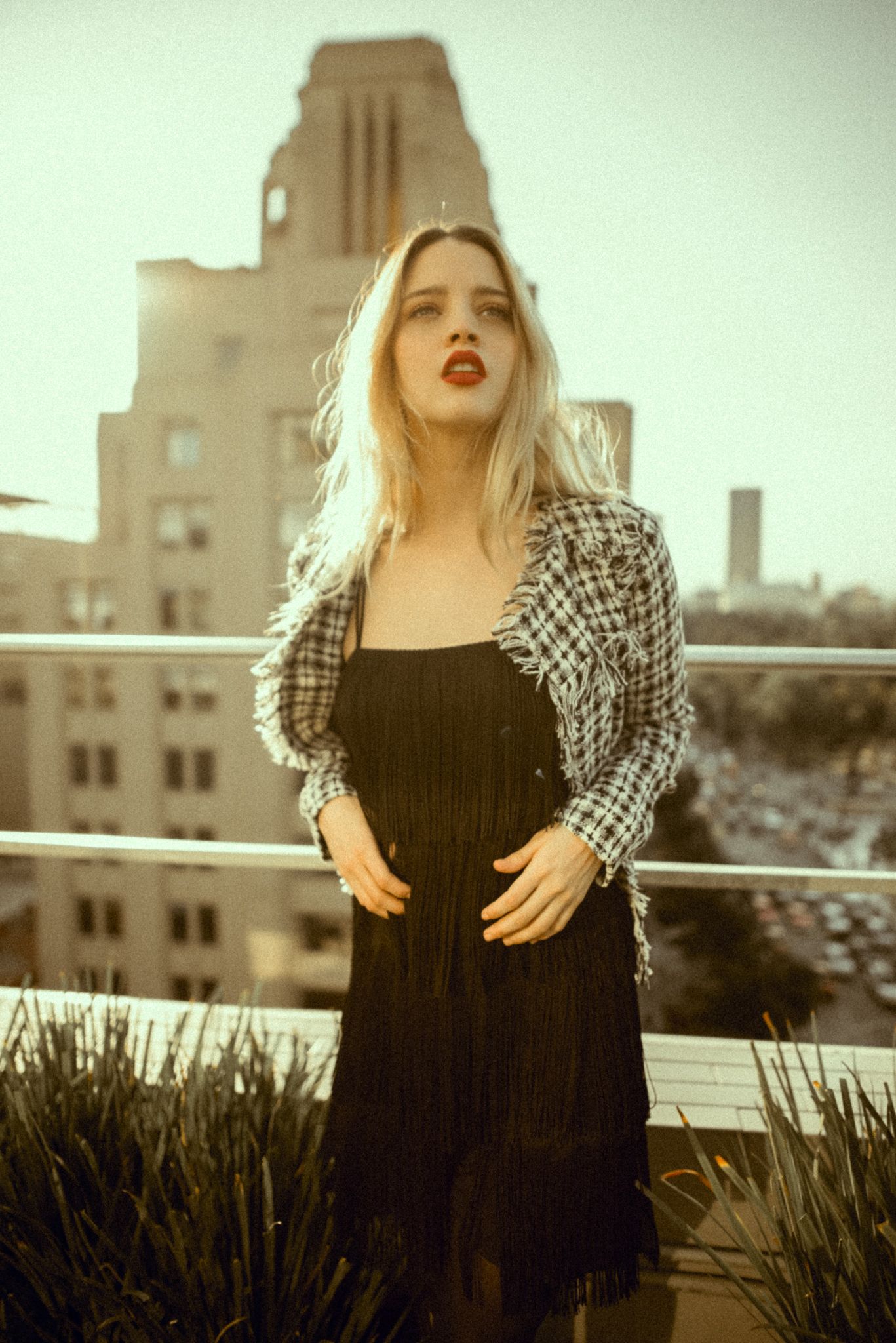 Rooftop fashion portrait in black dress and plaid jacket with city architecture background.