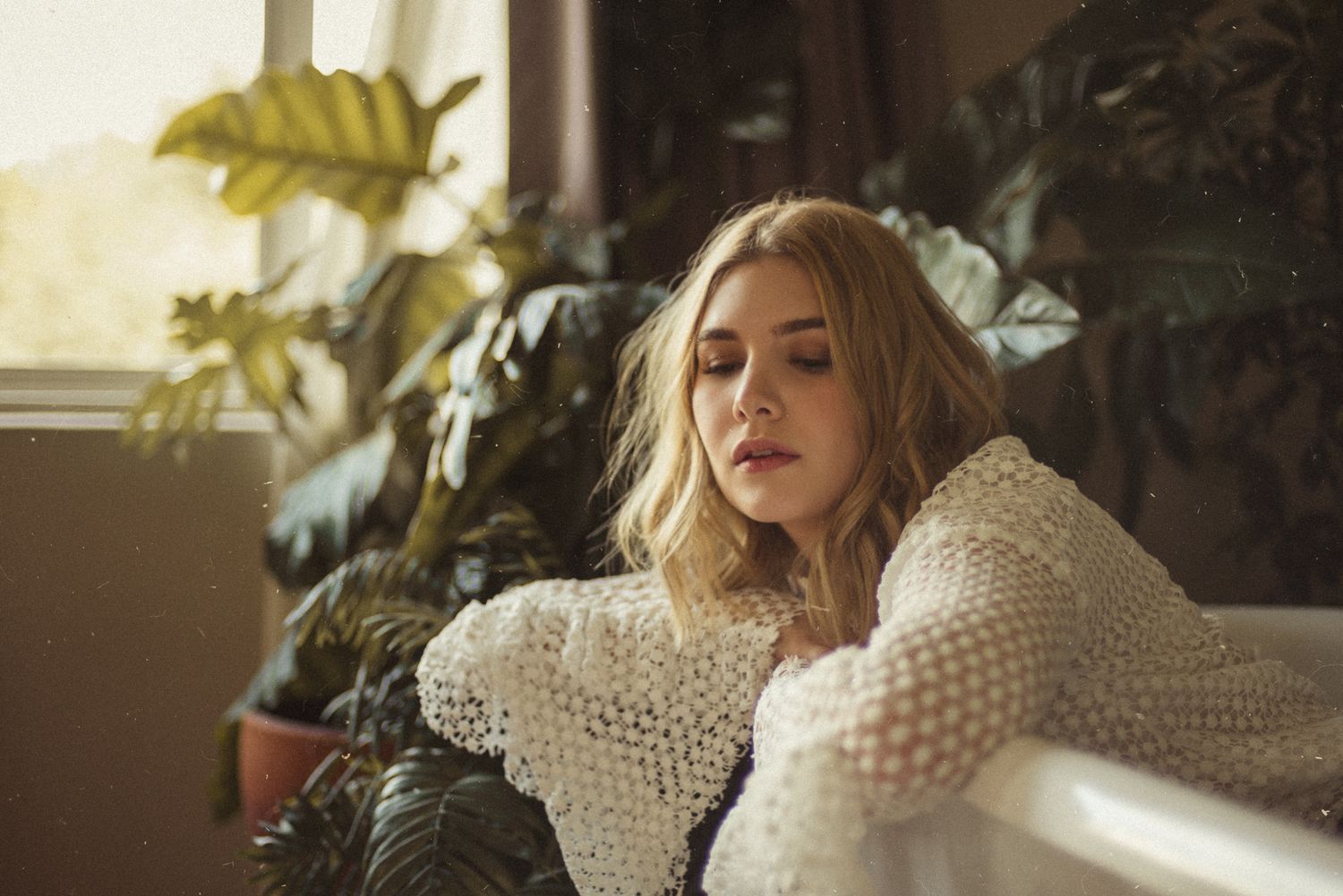 Woman in white knit sweater lounging near window with houseplants.