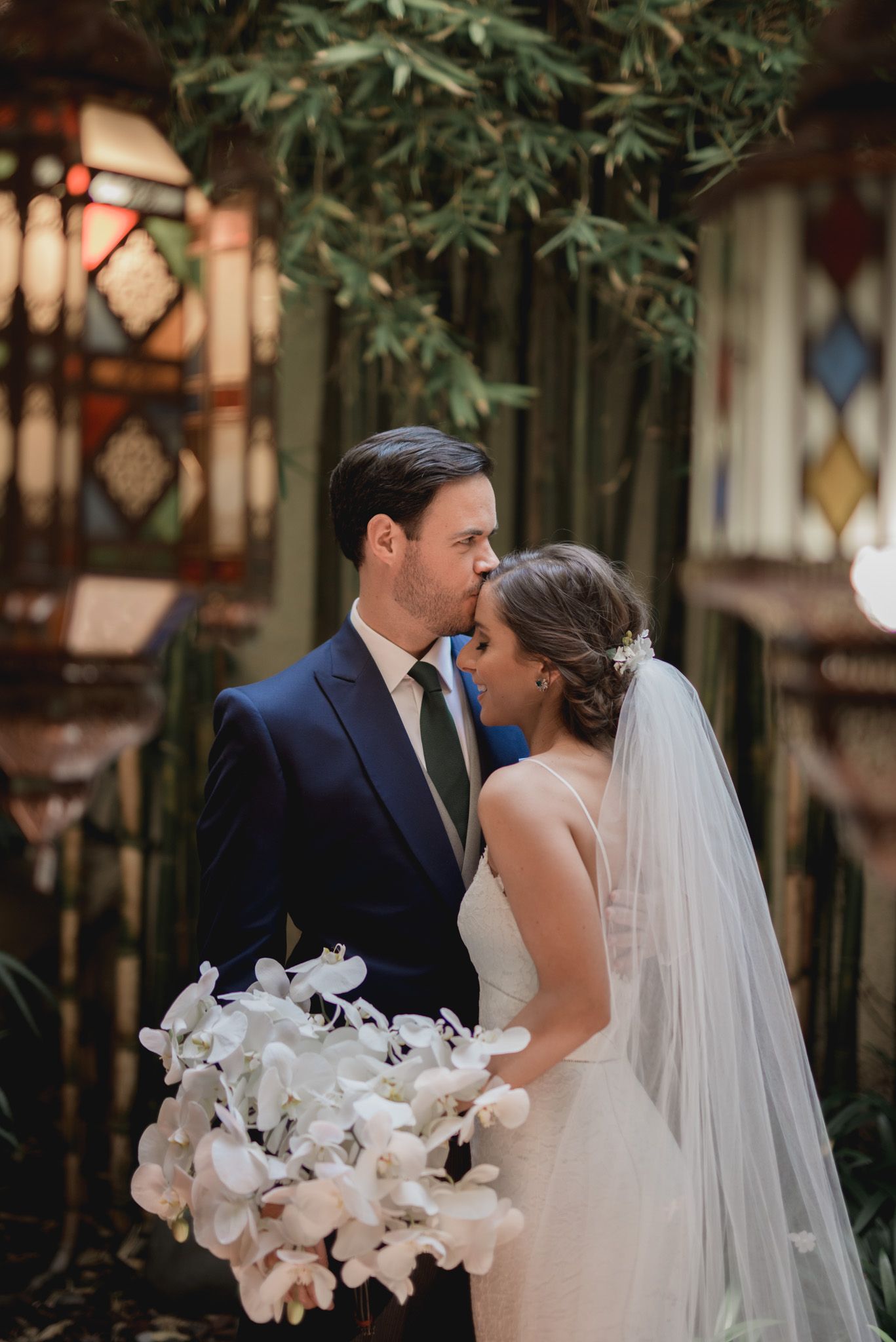Elegant wedding couple shares intimate moment in garden setting with white orchid bouquet and flowing veil.
