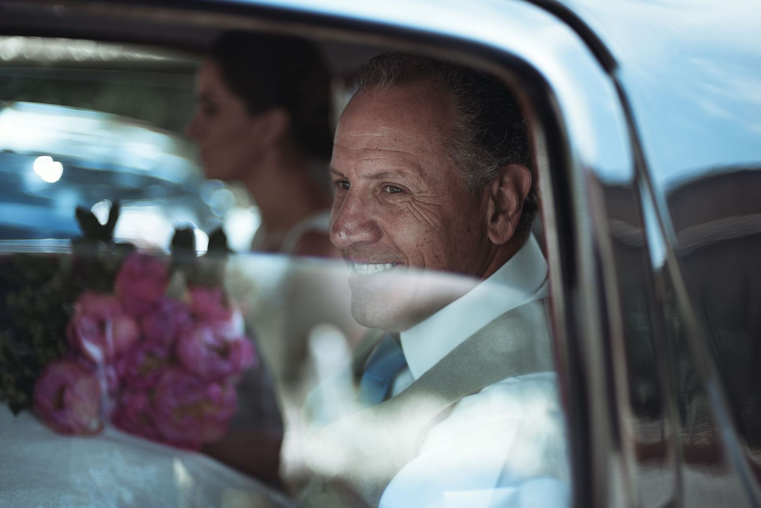 A person in formal white attire sits in a classic car with pink flowers visible through the window.