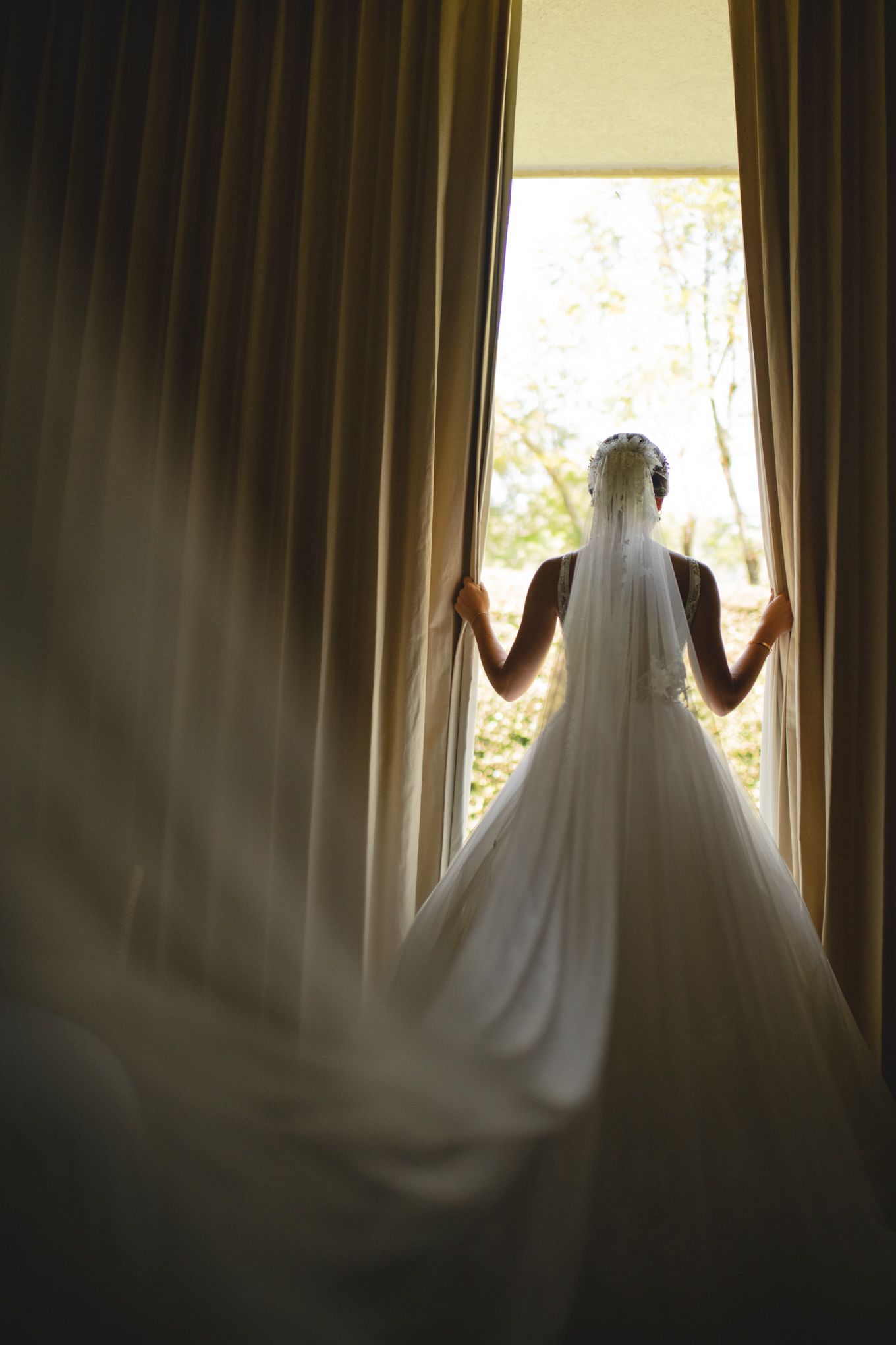Silhouette of bride in wedding dress and veil standing between curtains by window in dramatic lighting.