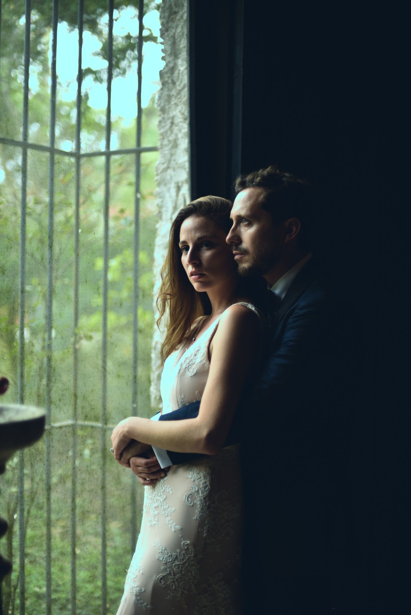 A romantic couple stands by a window in formal attire, with natural light filtering through trees outside.