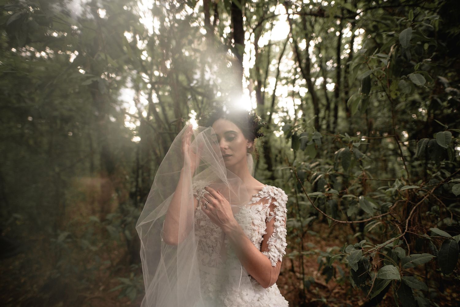 A bride in a lace wedding dress and veil poses ethereally in a misty forest during golden hour.