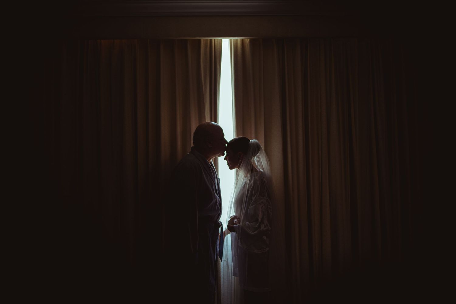 A silhouetted couple stands together in front of curtains with dramatic backlighting creating a romantic mood.
