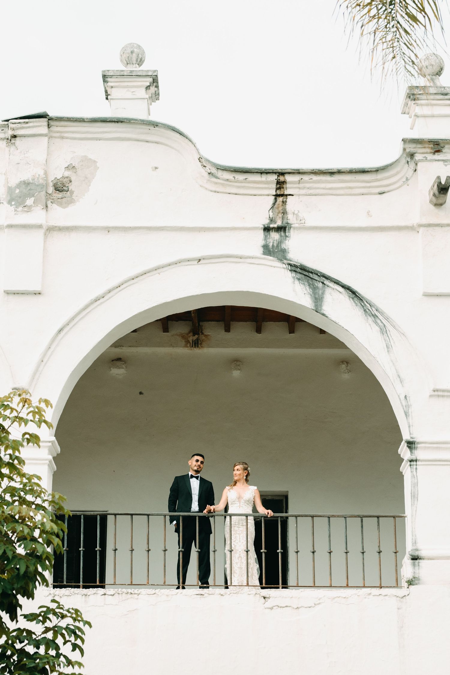 A couple stands on a white arched balcony of a Spanish colonial style building during a wedding photoshoot.