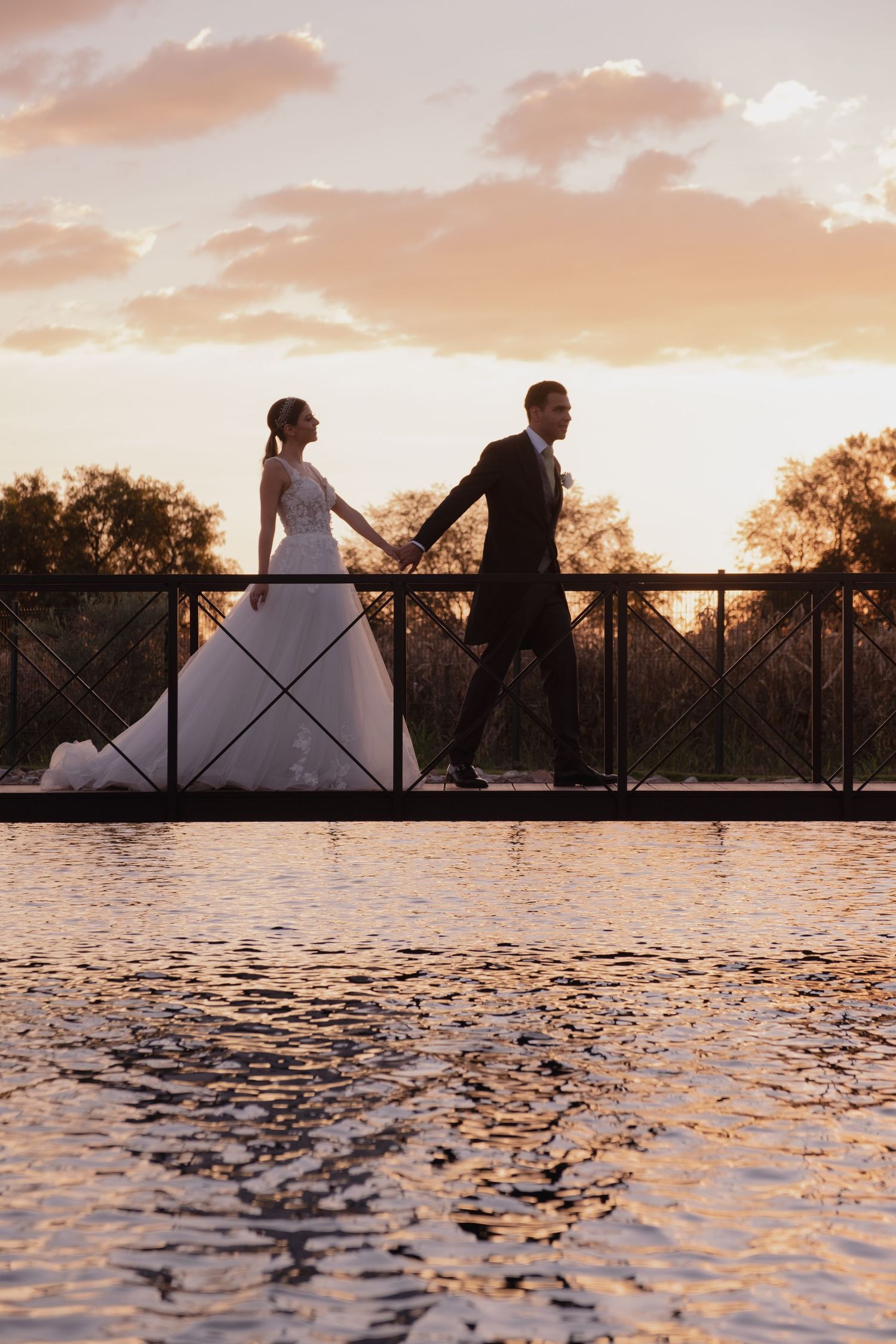 Silhouetted couple walking across a bridge during sunset with water reflections below.