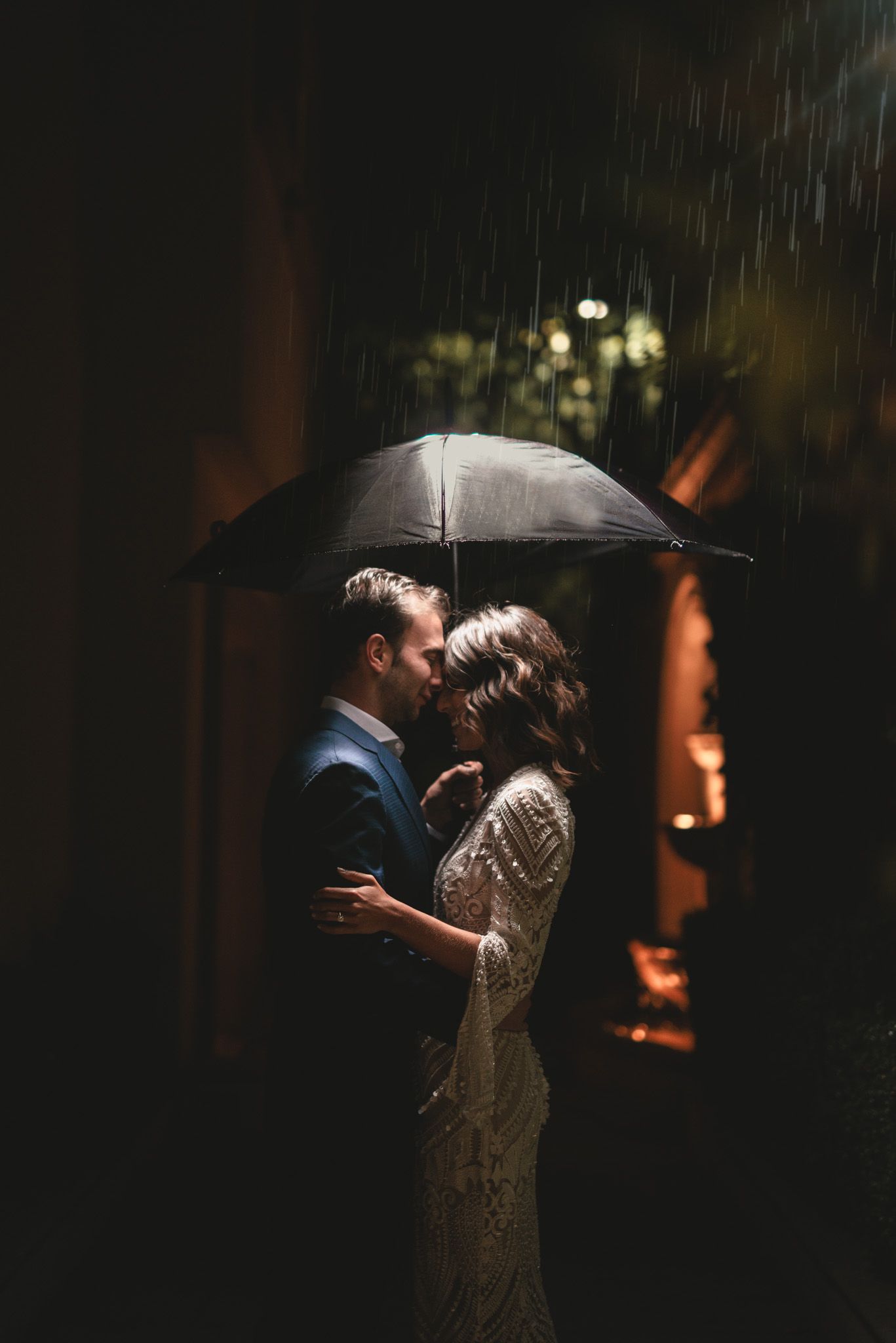 A romantic couple shares an intimate moment under an umbrella in the rain with dramatic moody lighting.