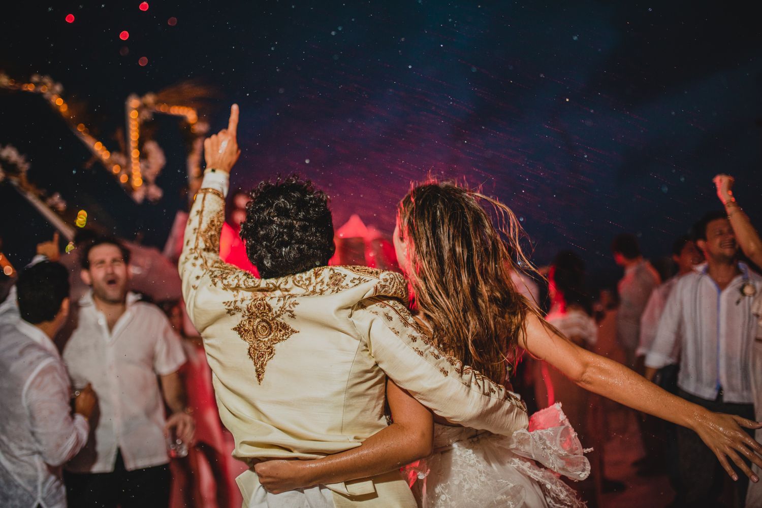 Wedding couple dancing together at reception with guests on dance floor under colorful nightclub lighting.