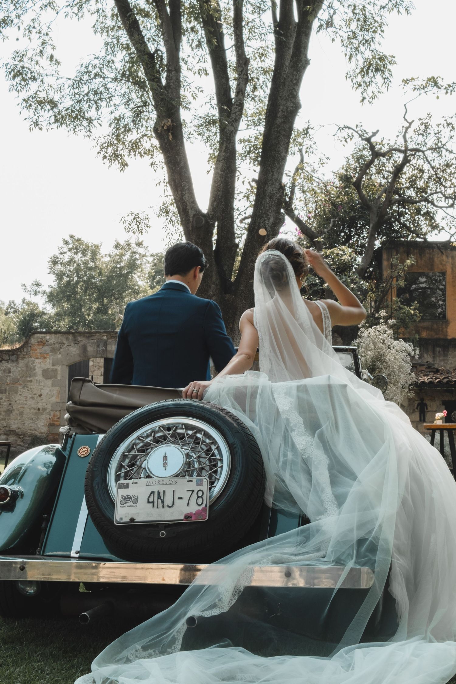 Newlyweds sitting in vintage car surrounded by flowing white veil and garden setting.